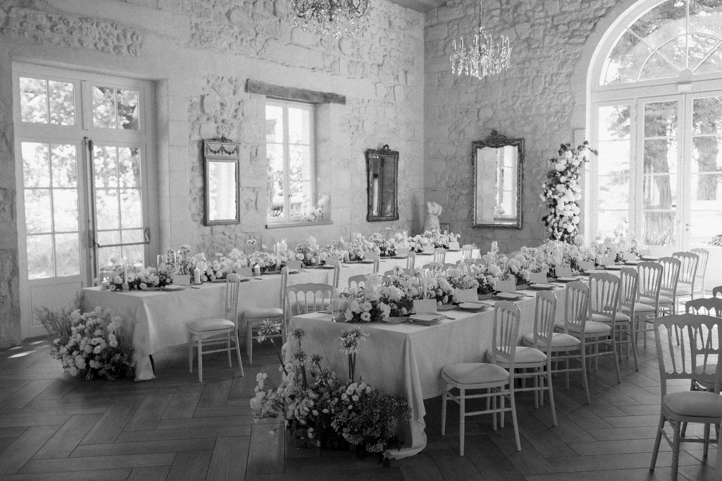 A wedding reception setup with long tables decorated with flowers and candles, surrounded by chairs, inside a room with stone walls, large windows, and chandeliers.
