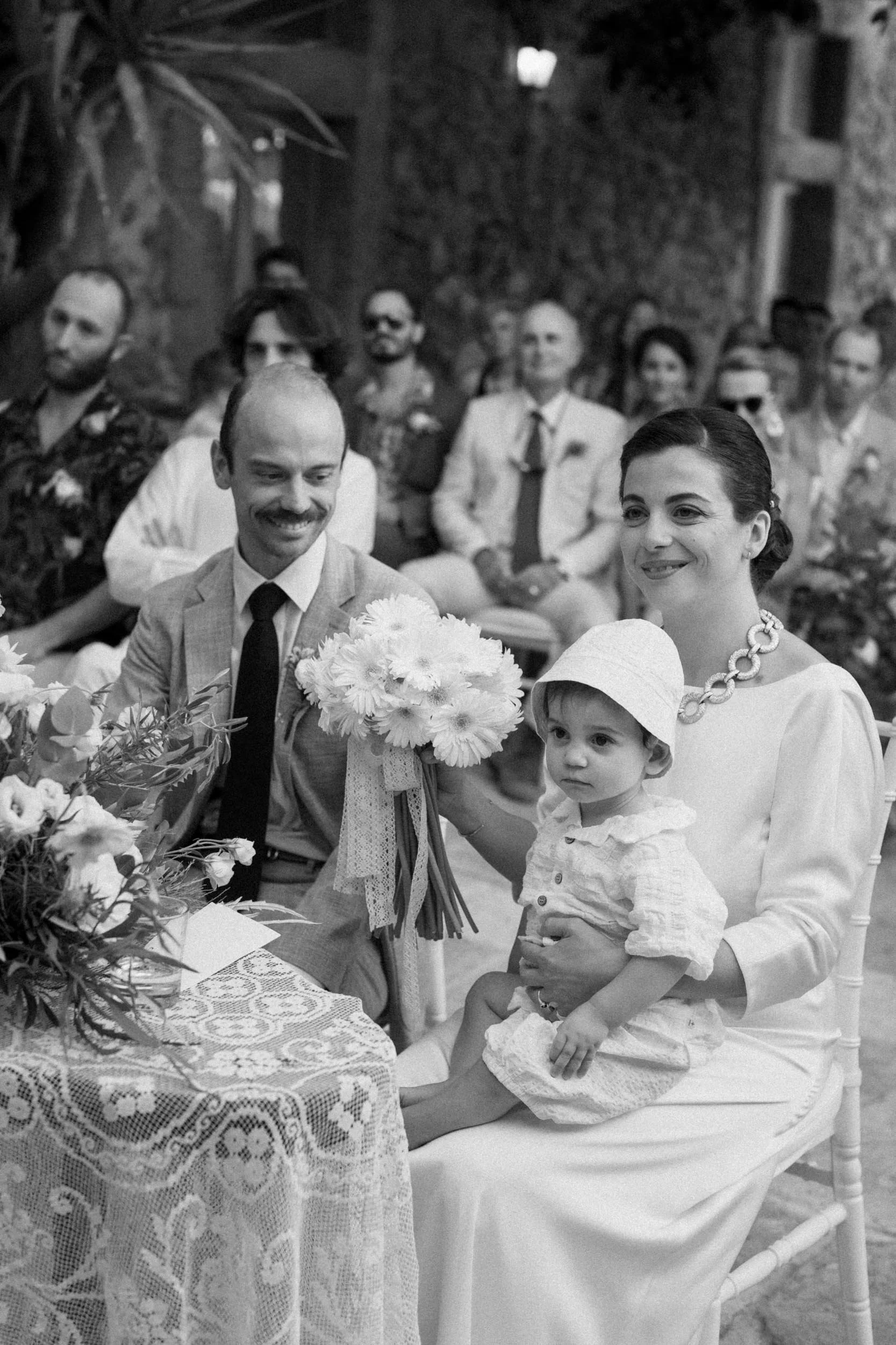 A woman holding a young girl sitting on her lap during a wedding ceremony, with a man next to them holding a bouquet of flowers. Guest are seated behind them.