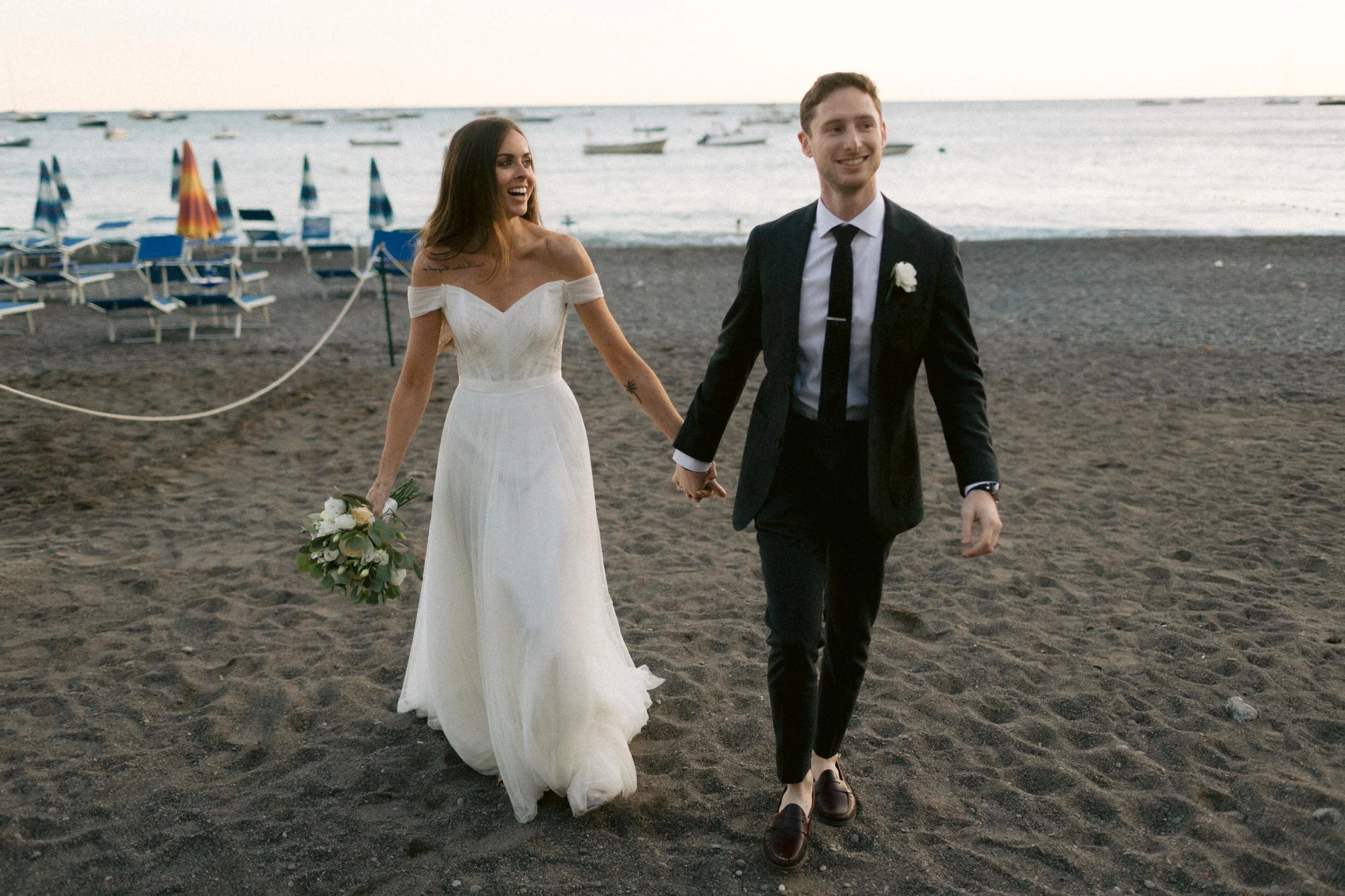 A newlywed couple walking hand in hand on a dark sandy beach, with boats and umbrellas visible in the background.