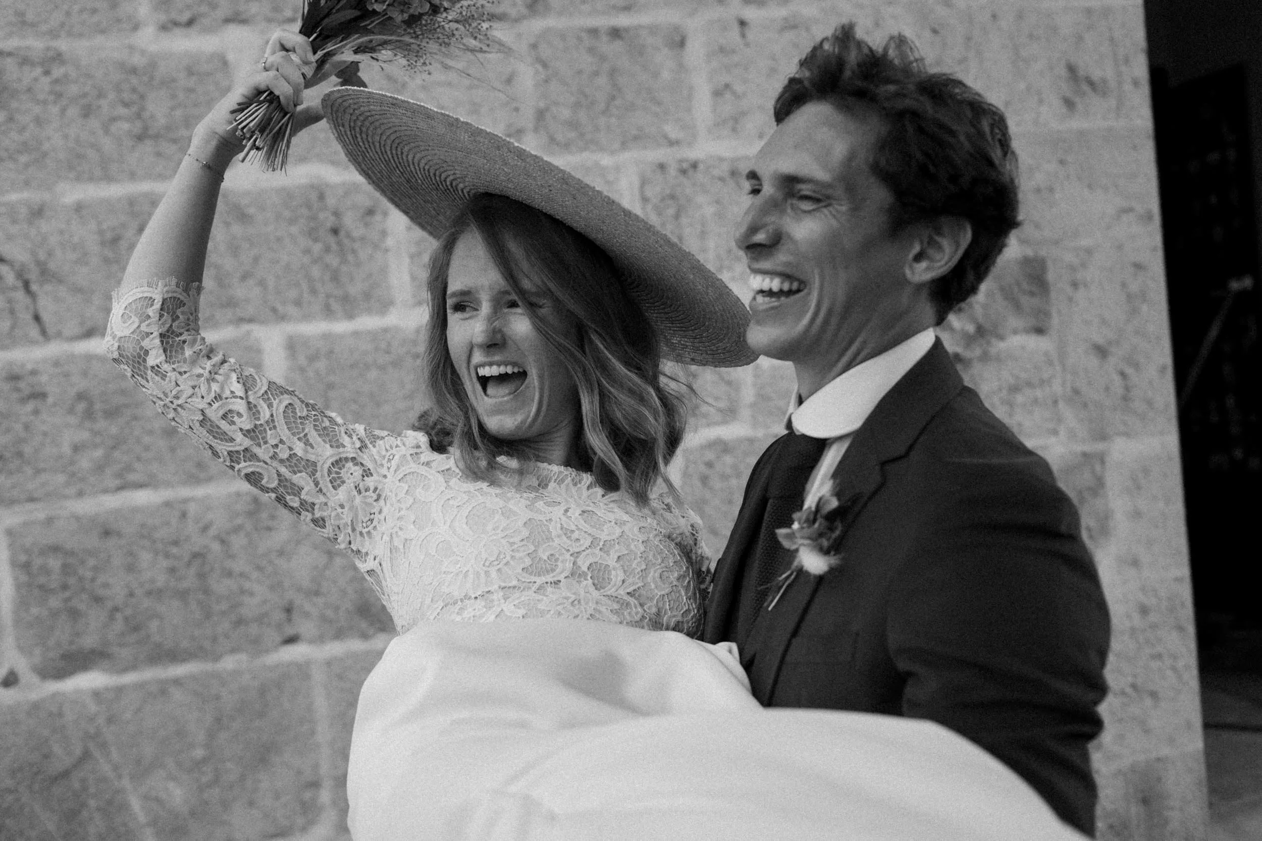 A man in a suit holding a smiling woman in a lace dress and large hat, against a brick wall.