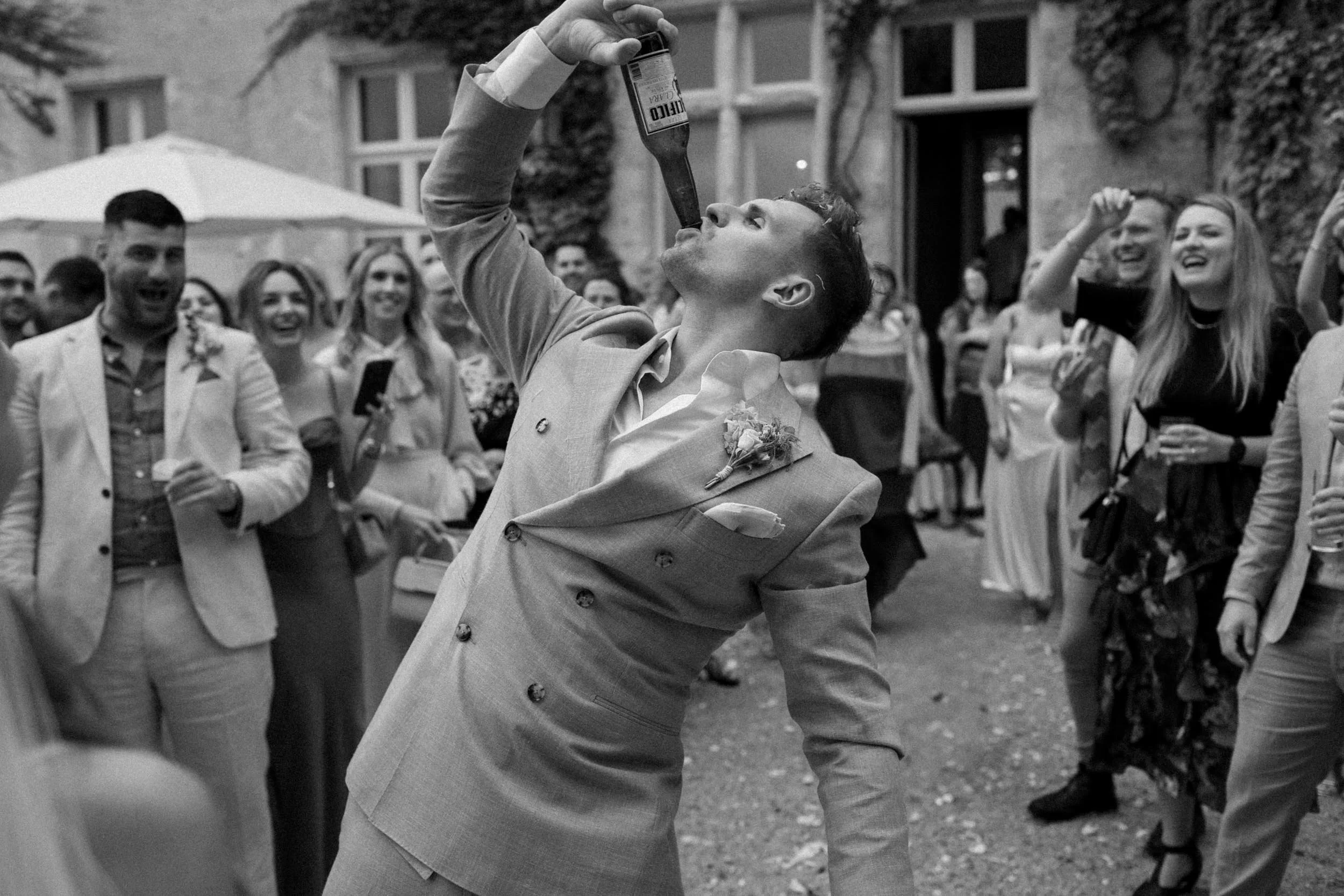 A man in a suit pouring a drink into his mouth at a wedding reception, surrounded by people dancing and celebrating outdoors.