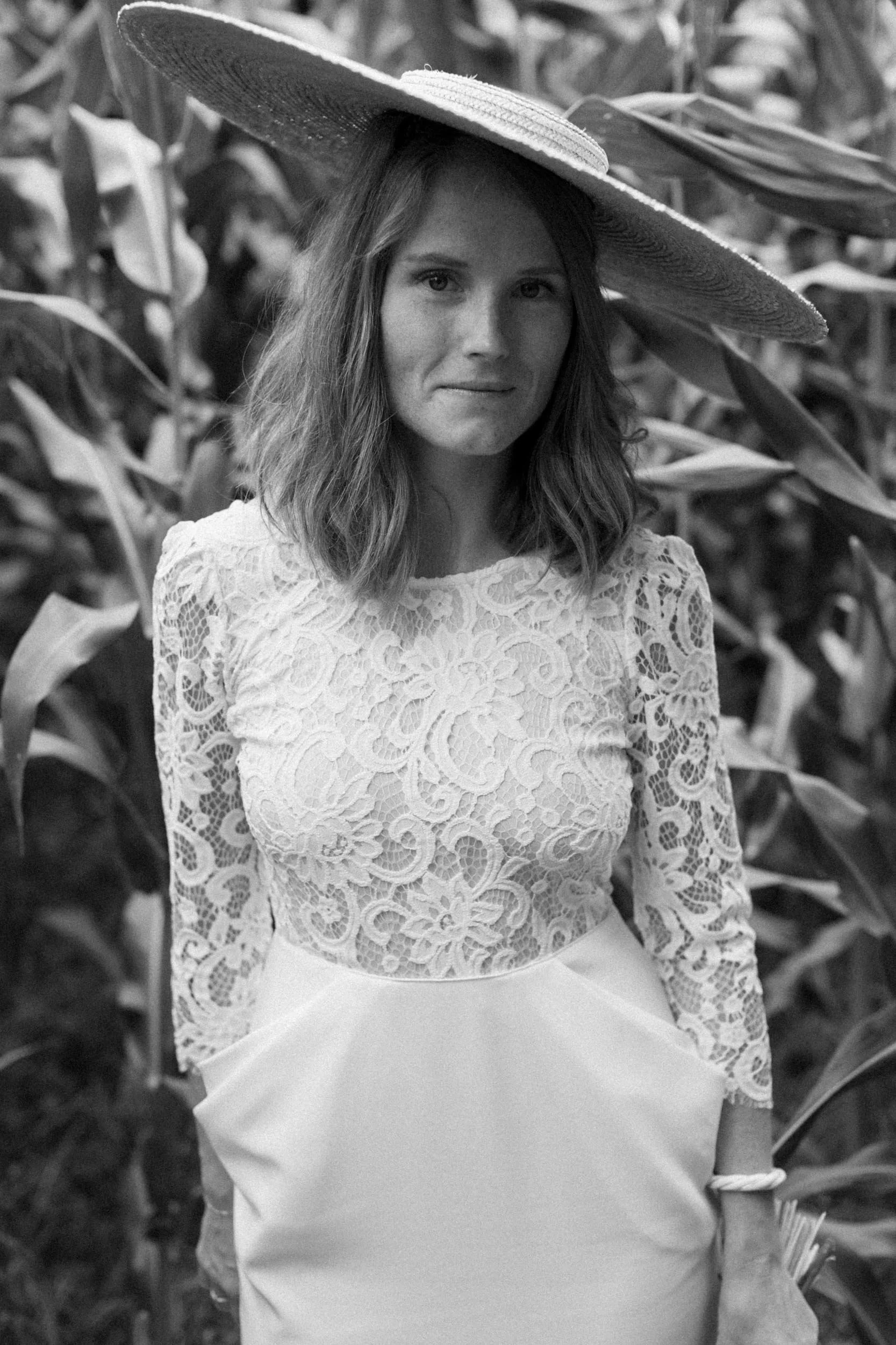 A woman in a floral lace top and a white skirt, wearing a large straw hat, standing amidst tall plants or cornfield.
