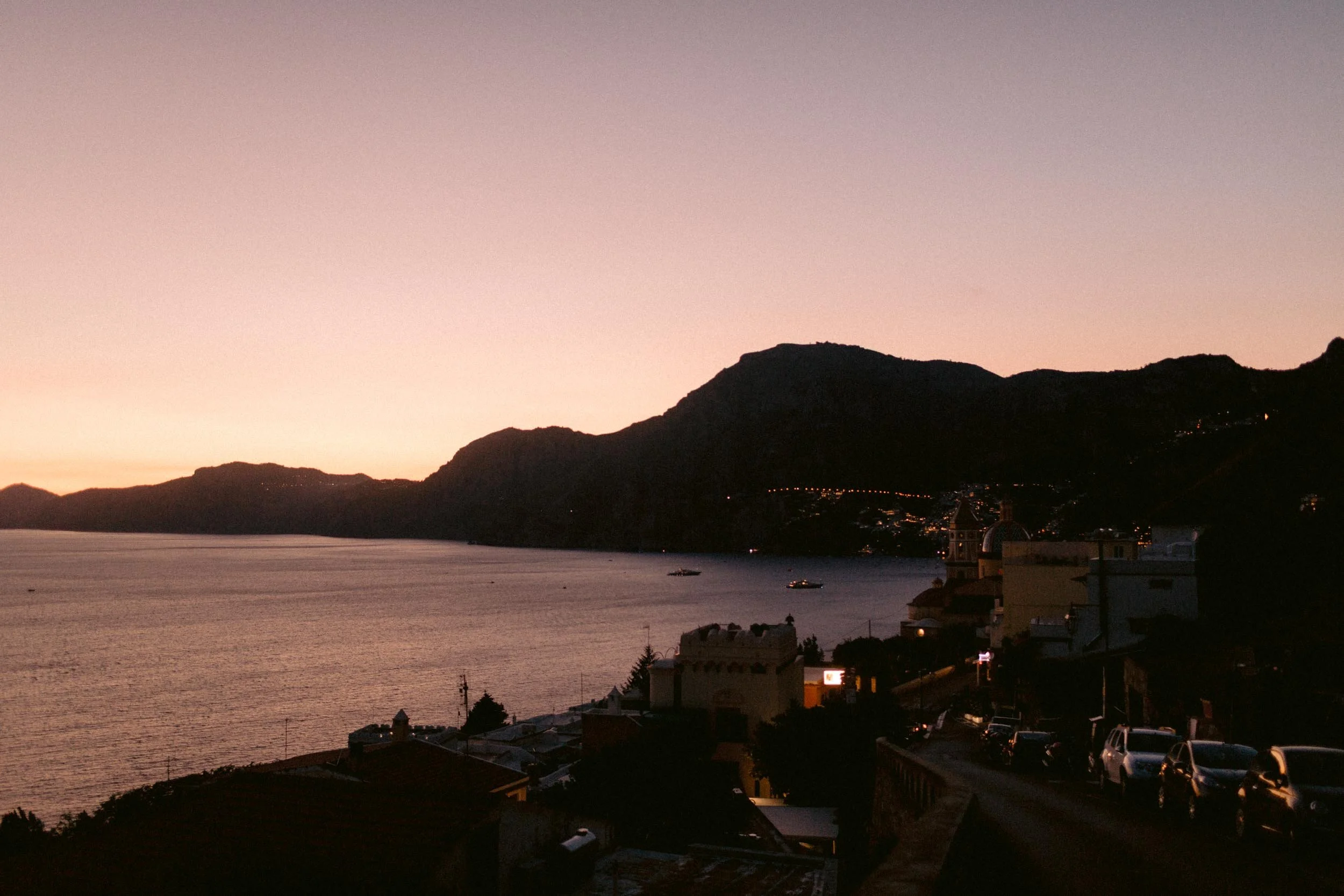 Sunset over a coastal town with mountains in the background and the water in the foreground. The sky has a gradient of pink and purple, with some buildings and cars visible in the darkened foreground.