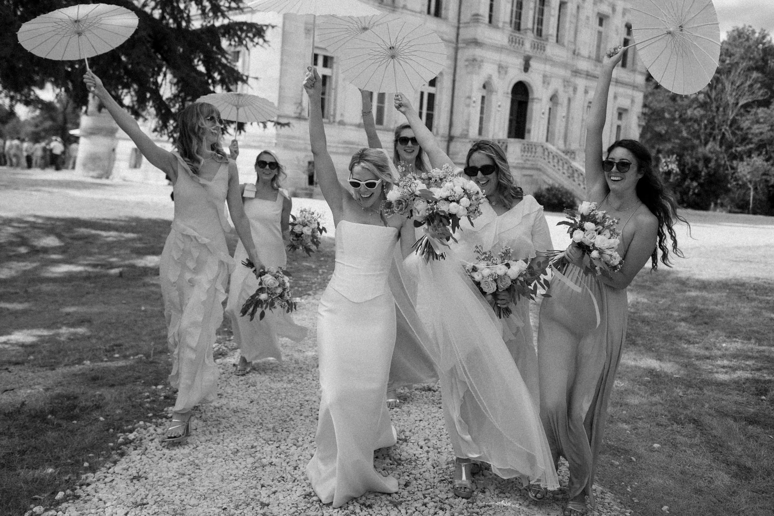 Group of women celebrating outdoors, holding bouquets and parasols, dressed in formal attire, in front of a historic building.