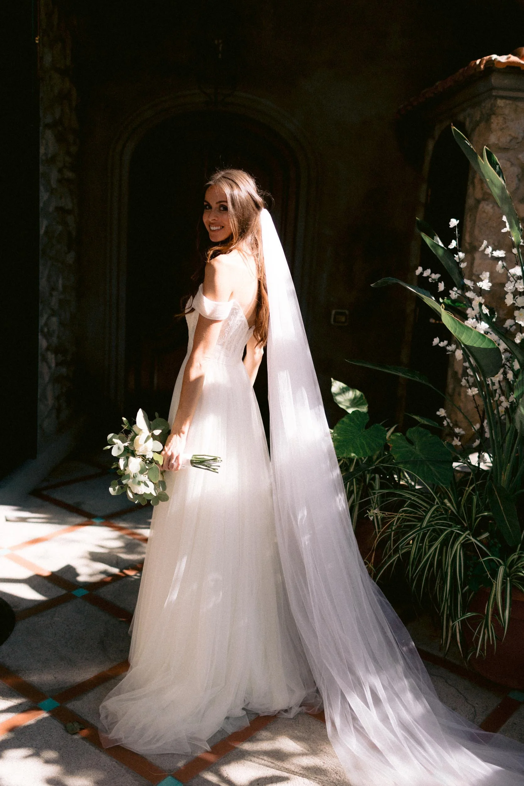 A bride in a wedding dress holding a bouquet, standing indoors near a stone wall and plants, smiling at the camera.