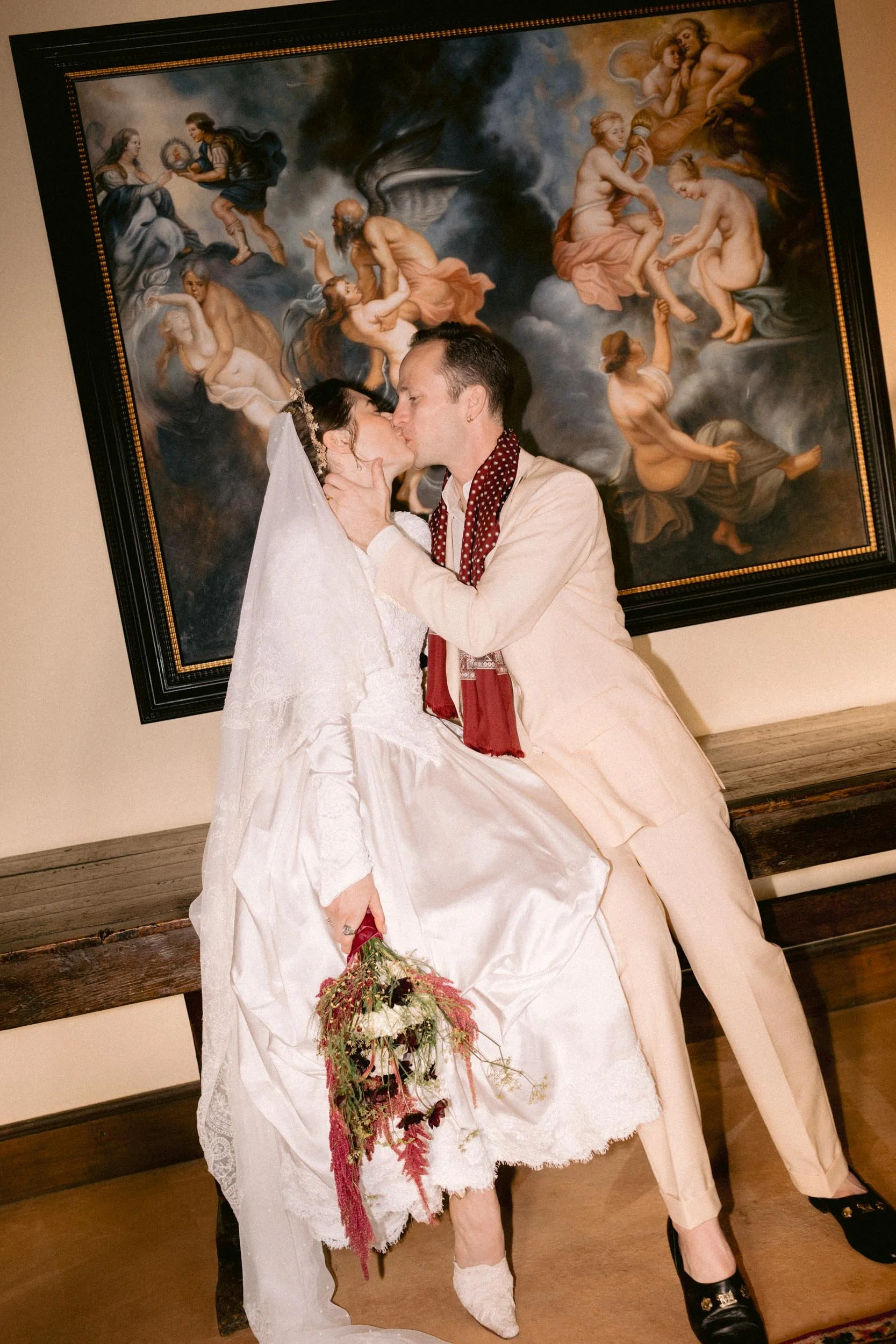 A couple is kissing in front of a large religious painting, with the bride dressed in a white wedding gown holding a bouquet, and the groom wearing a light-colored suit and dark shoes.