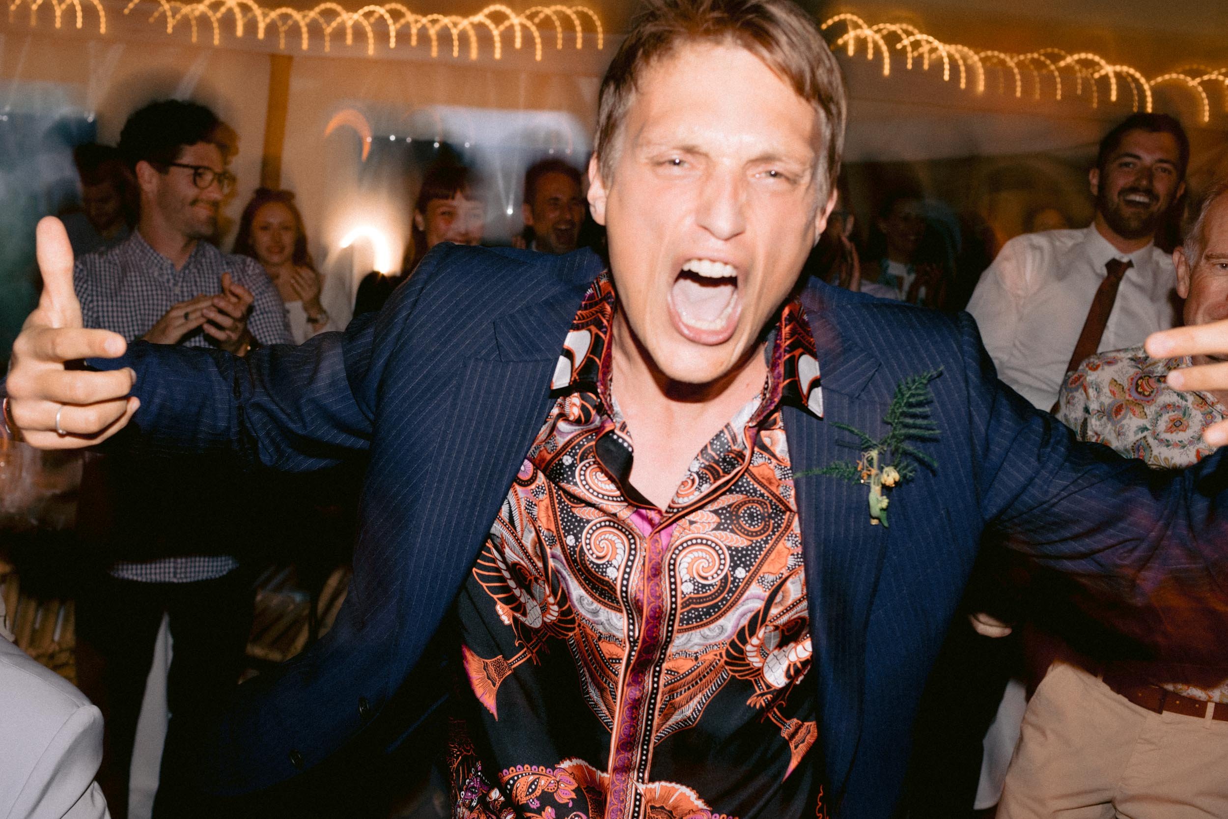 A man with short hair in a colorful, patterned shirt and dark blazer, celebrating with his arms outstretched and mouth open, at a party with people in the background.