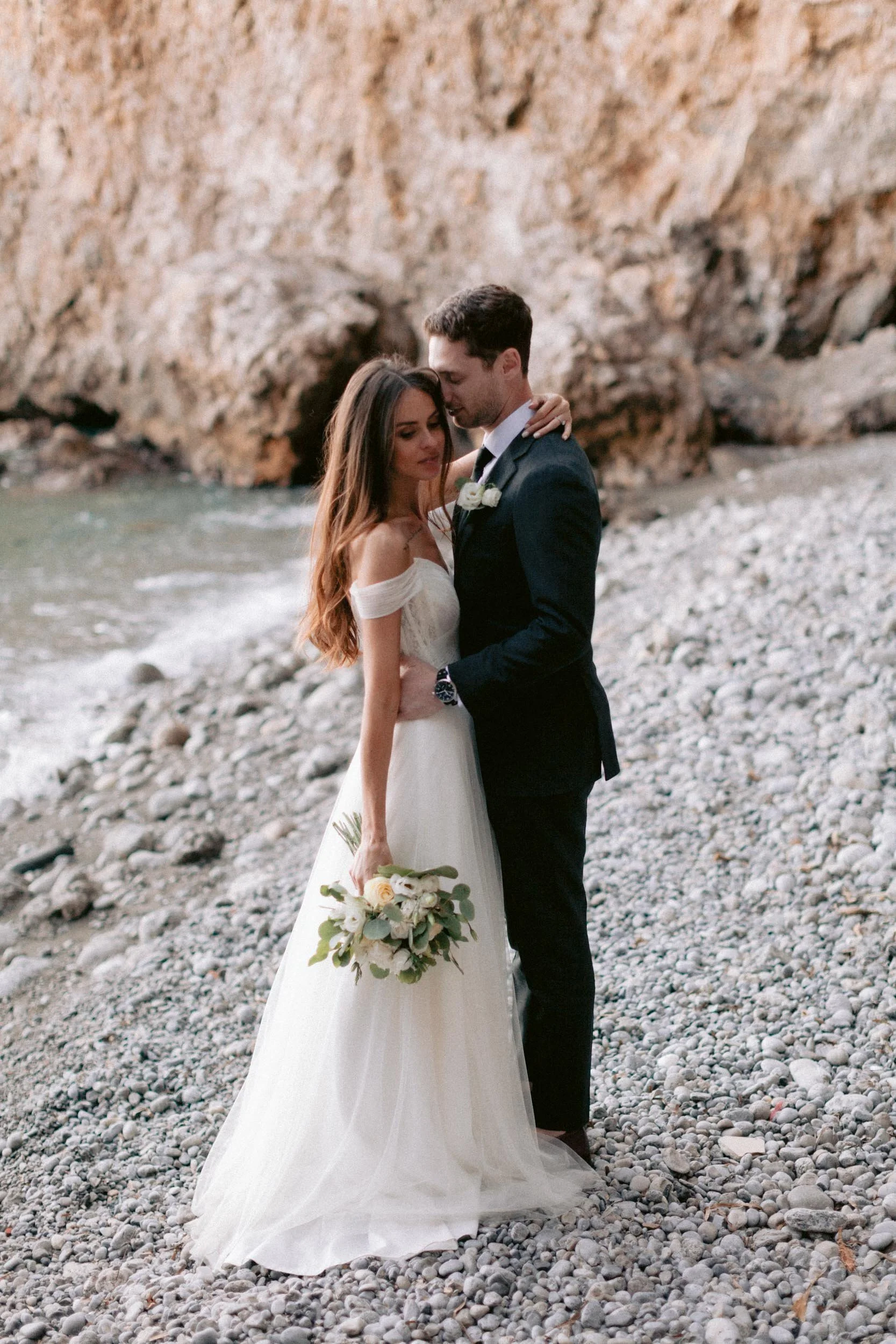 A newlywed couple embraces by the rocky shoreline with a large rock formation in the background. The bride is holding a bouquet of white flowers and wears a white off-shoulder wedding dress. The groom wears a dark suit with a white shirt, black tie, 