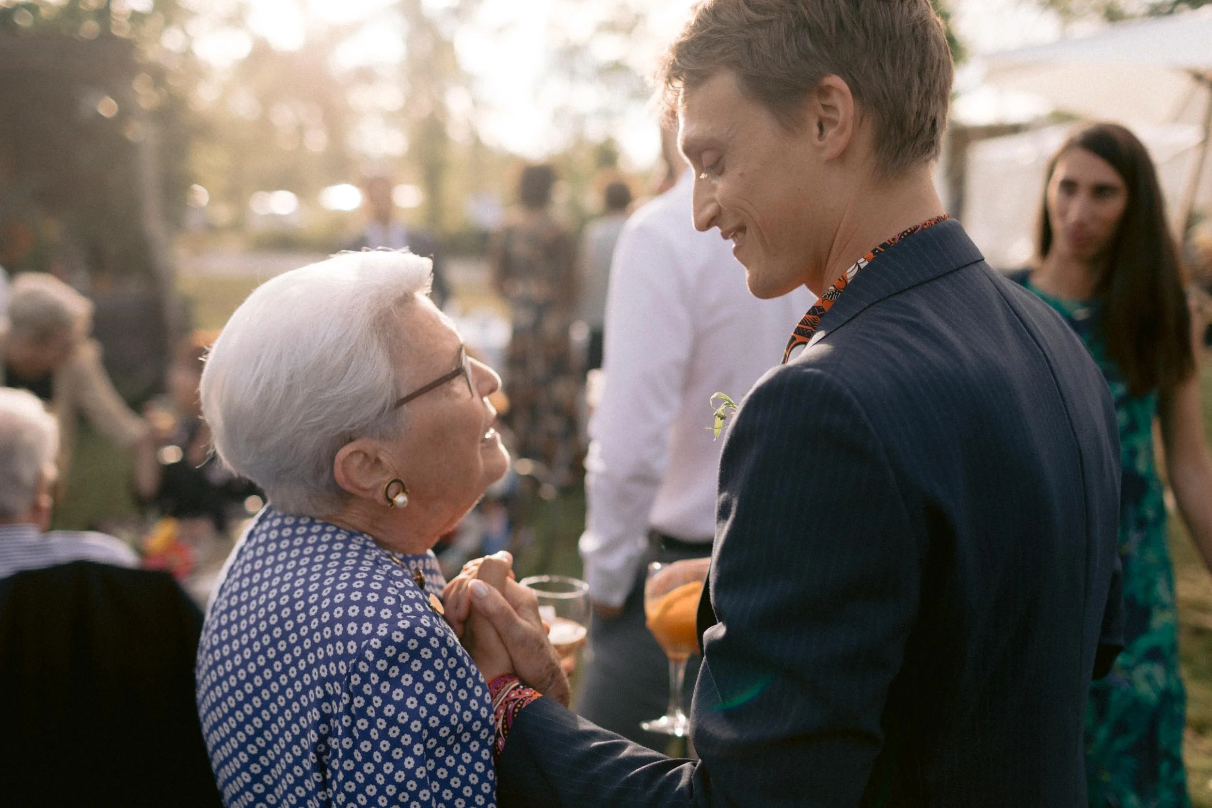 An elderly woman and a man are smiling and holding hands at an outdoor gathering during sunset, with other people in the background.