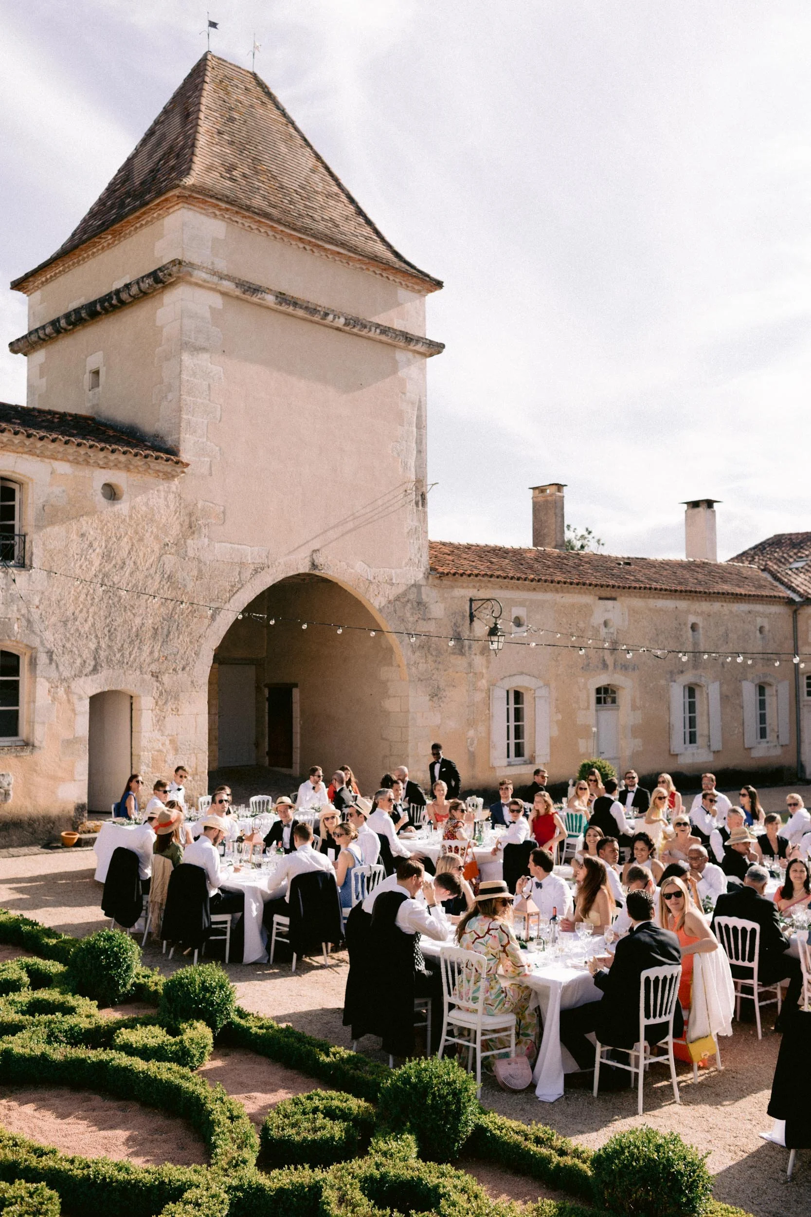 Outdoor wedding reception in a courtyard near a stone castle, with guests sitting at long tables decorated with white tablecloths and glasses, and string lights hanging overhead.