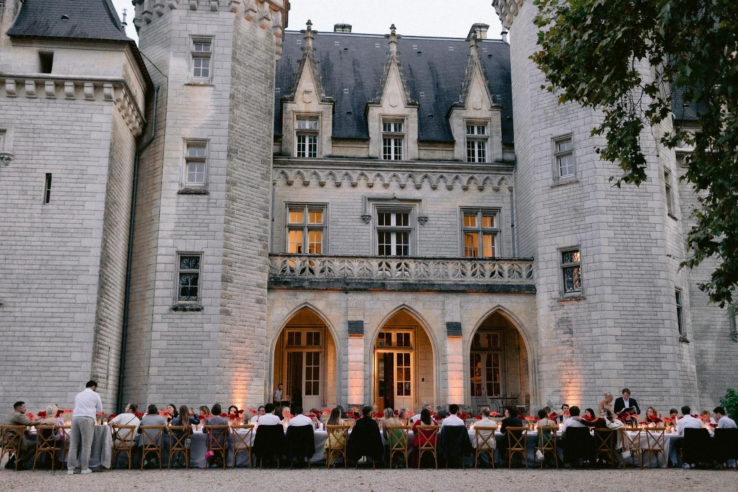 An outdoor dinner party at a banquet table set in front of a large historic castle with arched entrances and multiple windows, at dusk with warm lighting.