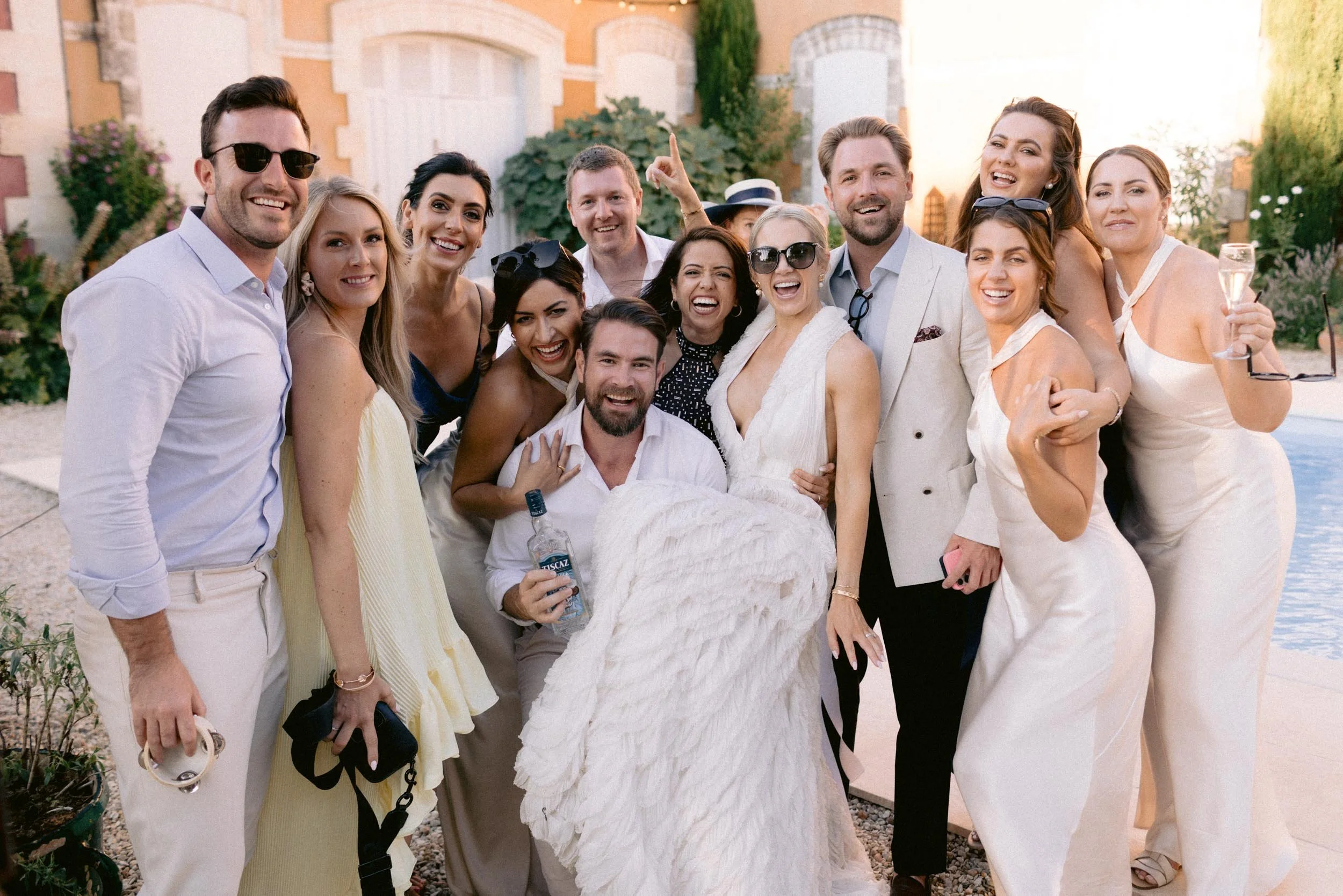 A group of people celebrating a wedding outdoors, with people smiling and posing for the camera, some wearing sunglasses and holding drinks.