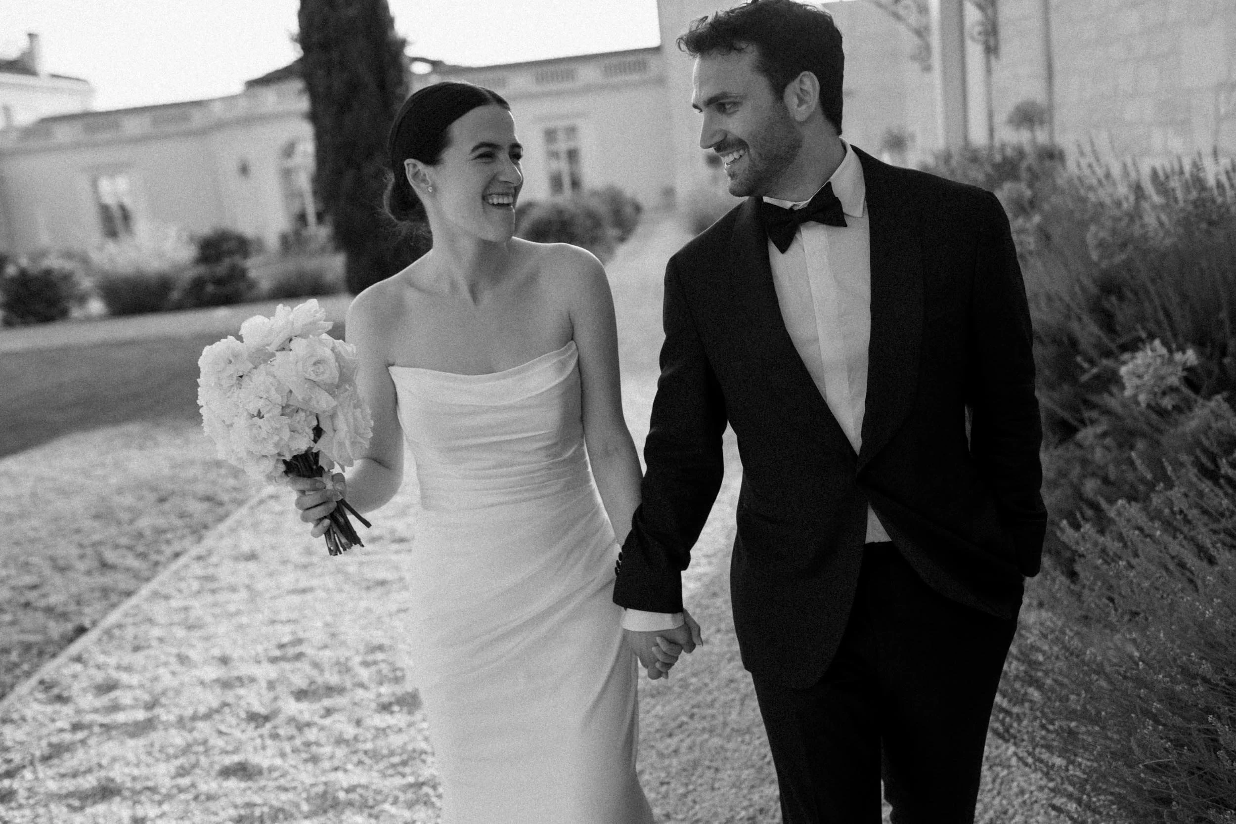 A black-and-white photo of a smiling bride and groom holding hands and walking outdoors, with the bride holding a bouquet of flowers.