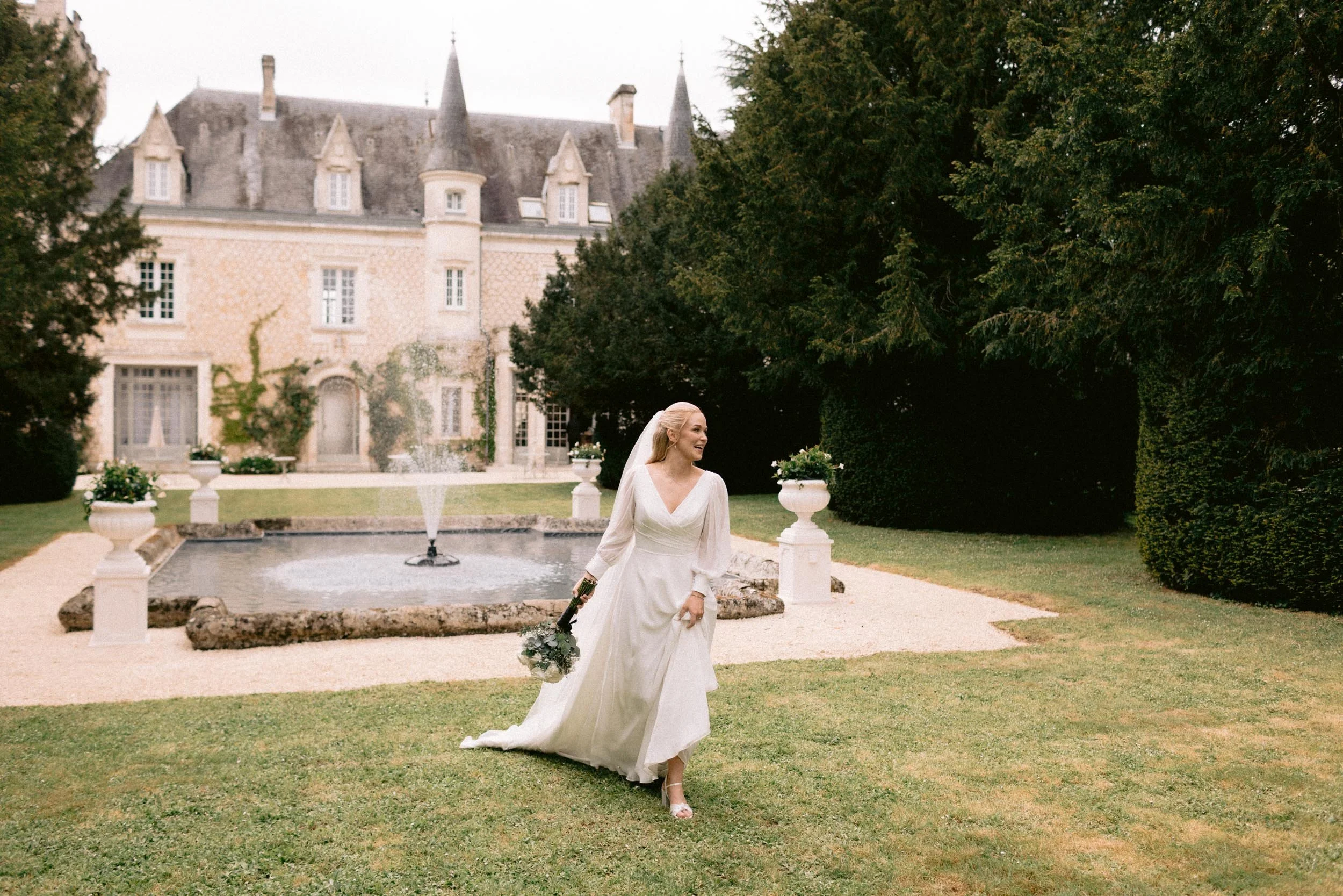 A bride in a white wedding dress holding a bouquet stands in a garden with a fountain behind her, a large castle-like building in the background, and trees surrounding the area.