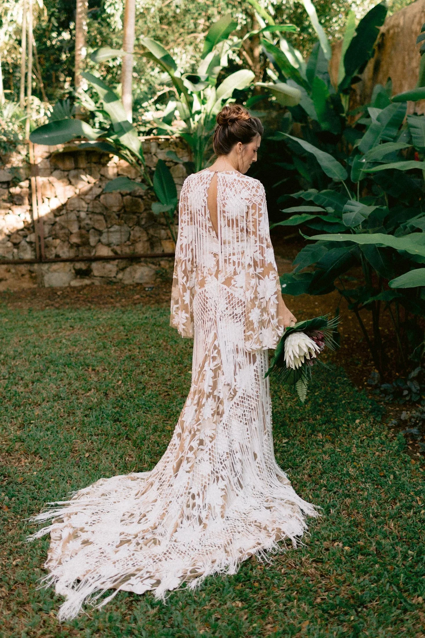 A woman in a long, intricate white lace dress holding a bouquet of flowers, standing in a lush garden with large green leaves and a stone wall in the background.