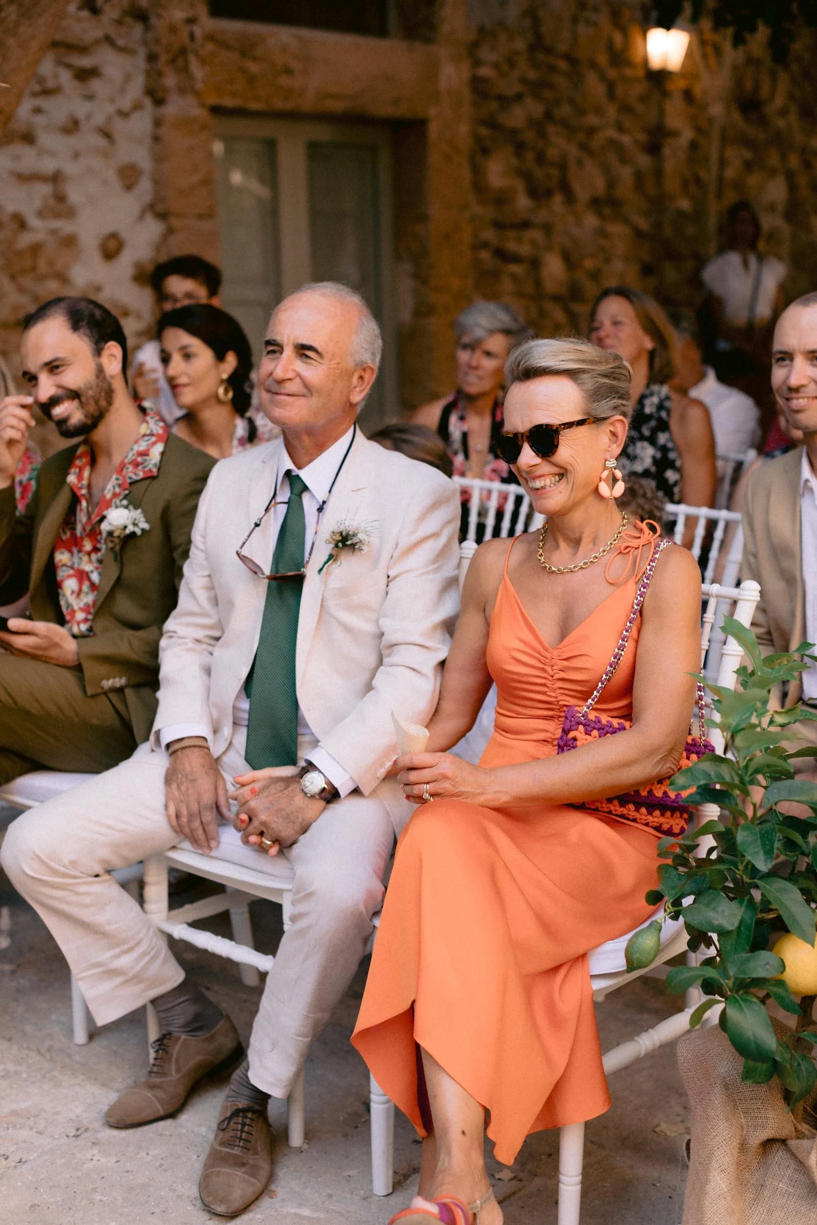 People sitting and smiling at an outdoor event, dressed in summer attire, with old stone walls in the background.