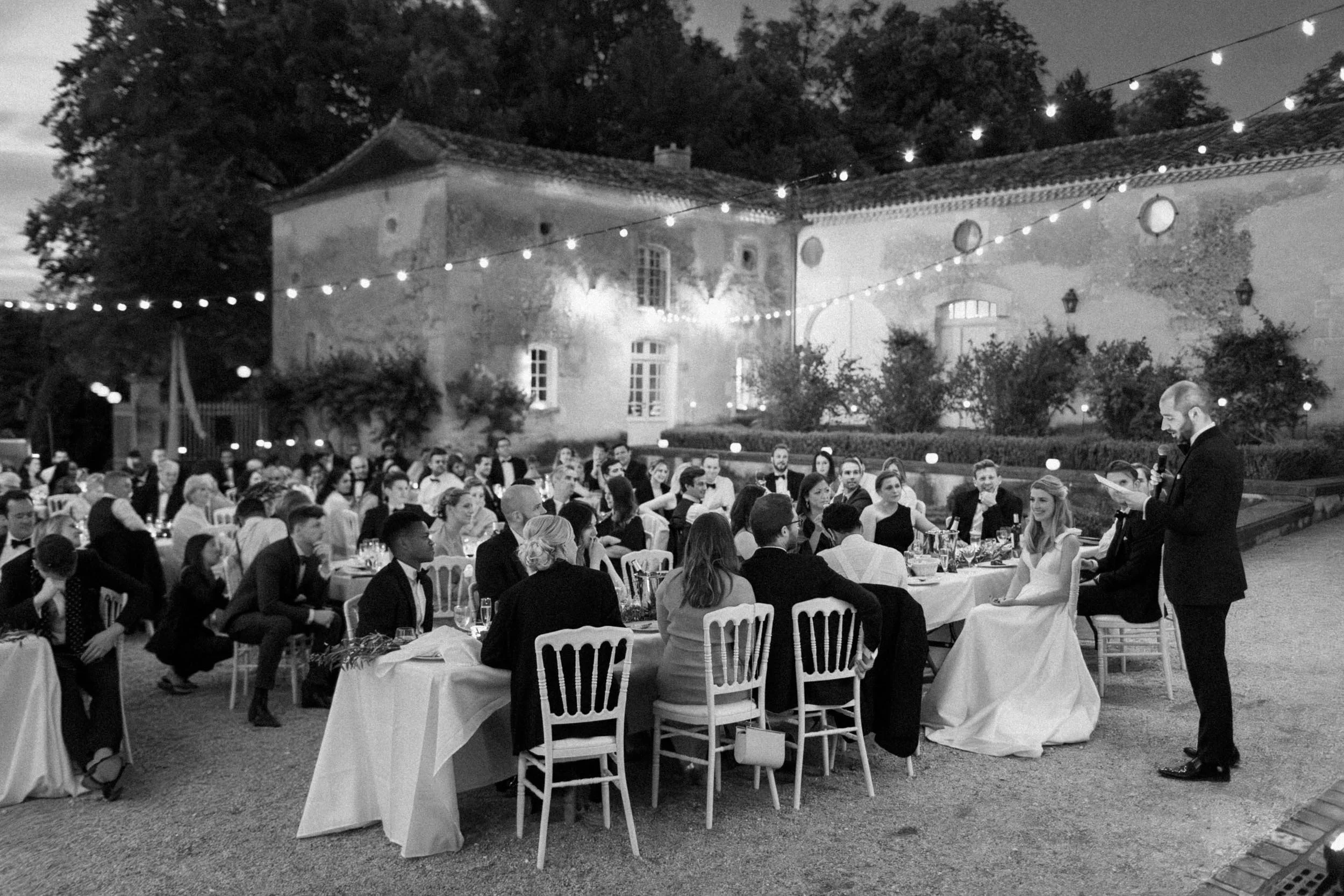 Black and white photo of an outdoor wedding reception under string lights at dusk or evening. Guests seated at long tables, with a bride and groom at the front, and a man reading or giving a speech.