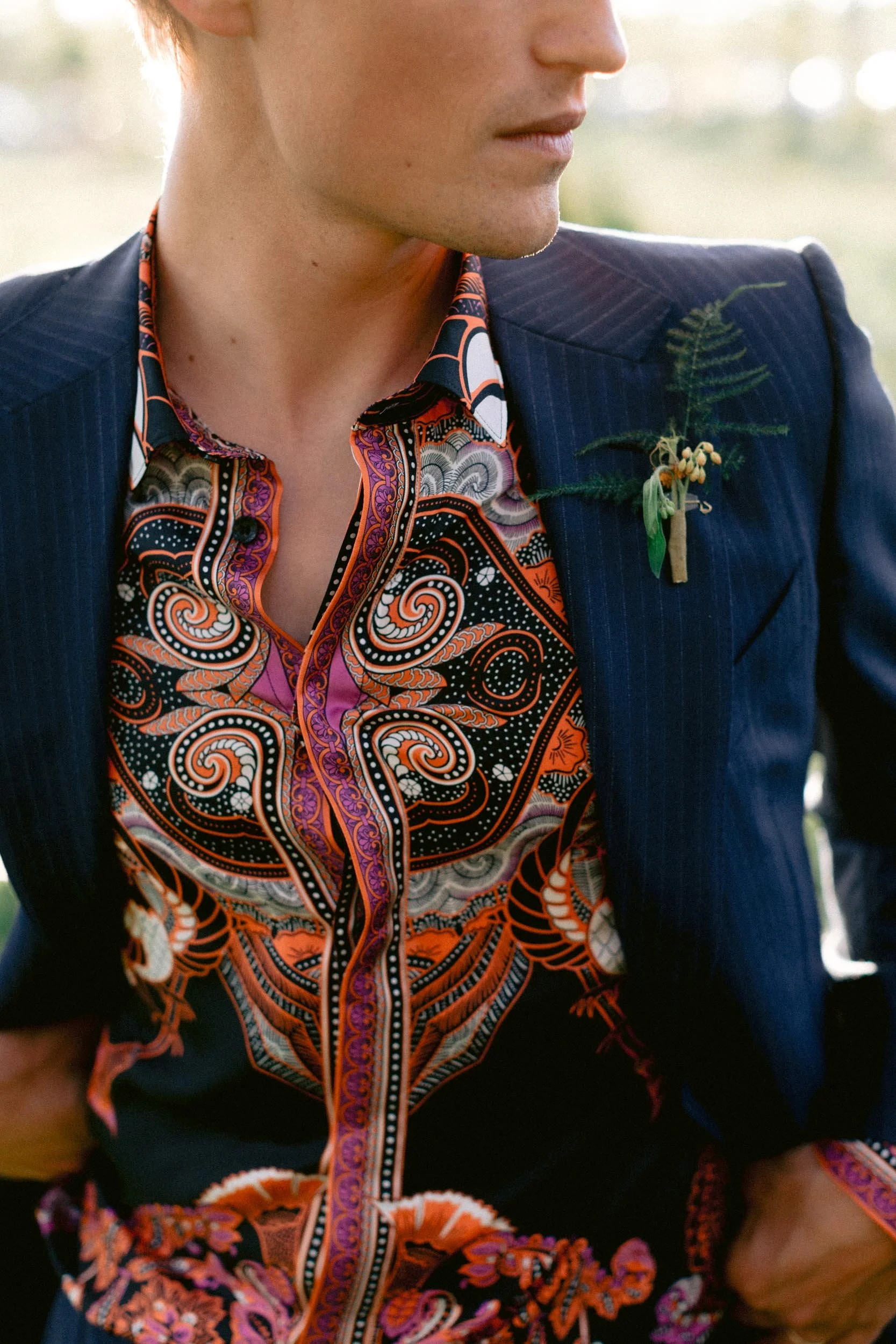 A person dressed in a dark pinstripe suit with a colorful paisley, floral, and swirl patterned shirt underneath. They have a small boutonniere with fern and small berries pinned to their blazer.