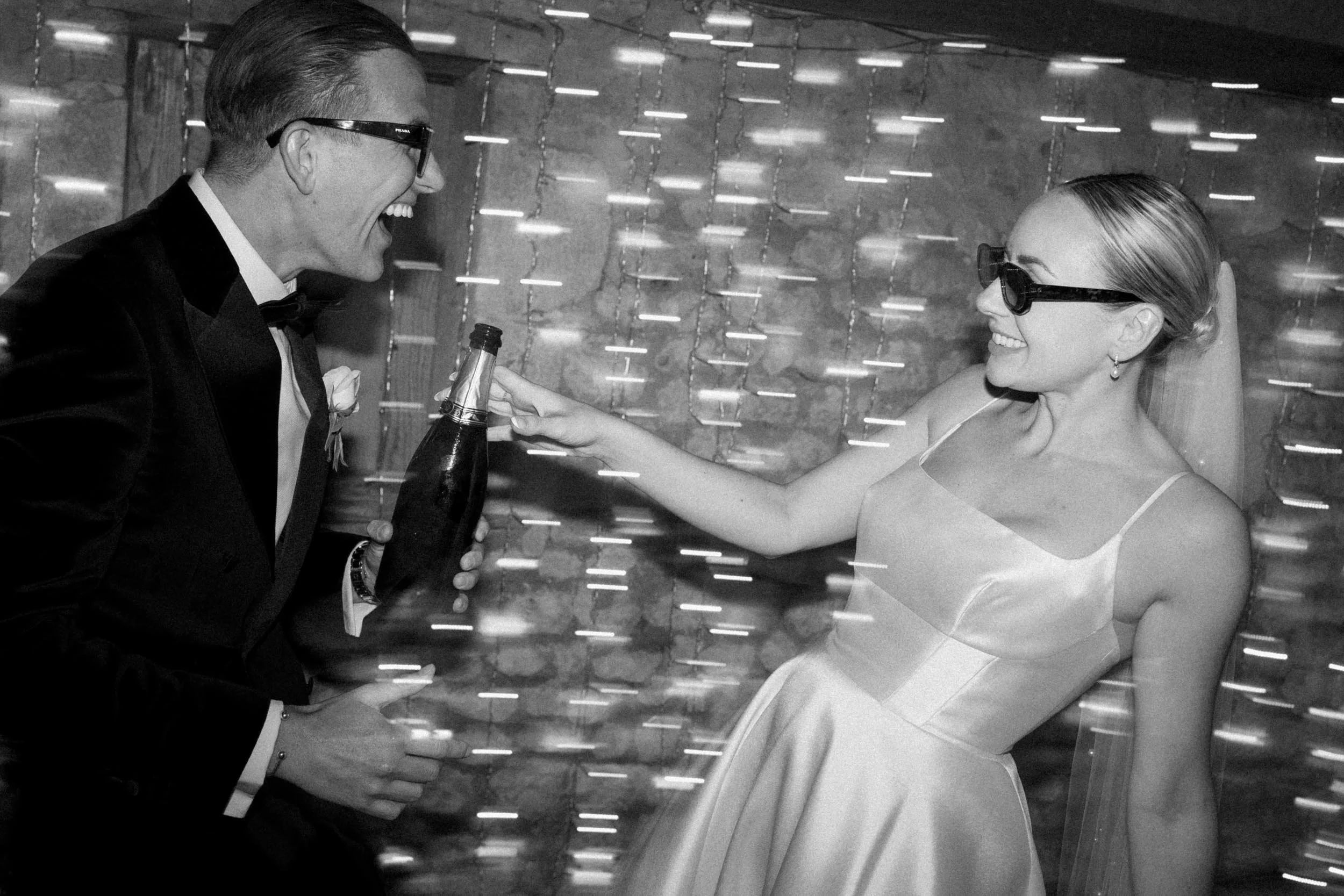A man in a tuxedo and a woman in a dress and sunglasses are sharing a joyful moment at a celebration, with the man holding a bottle of champagne and the woman reaching out to it.