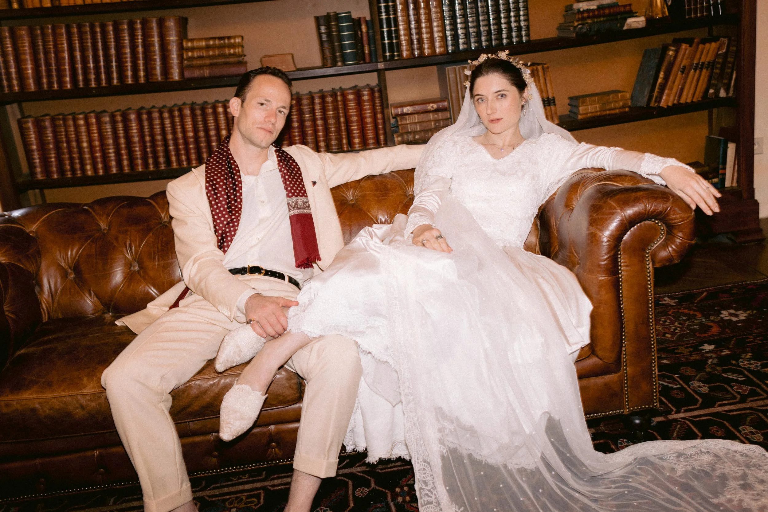 A couple sitting on a leather couch, with a woman in a white wedding dress and a man in light-colored suit and scarf. They are in a room with bookshelves in the background.