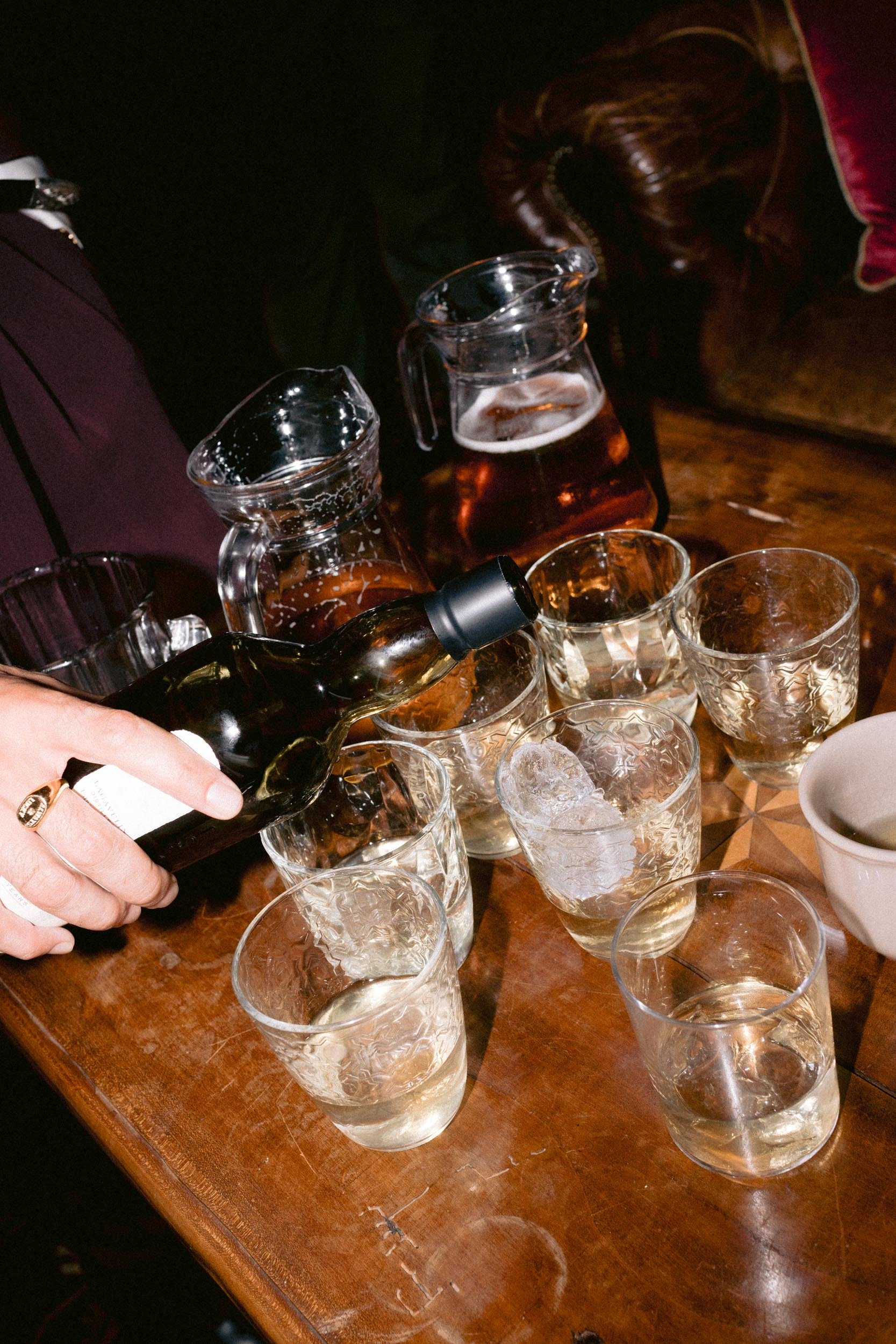 Person pouring whiskey into glasses with ice on a wooden table, surrounded by pitchers of water and other glasses.