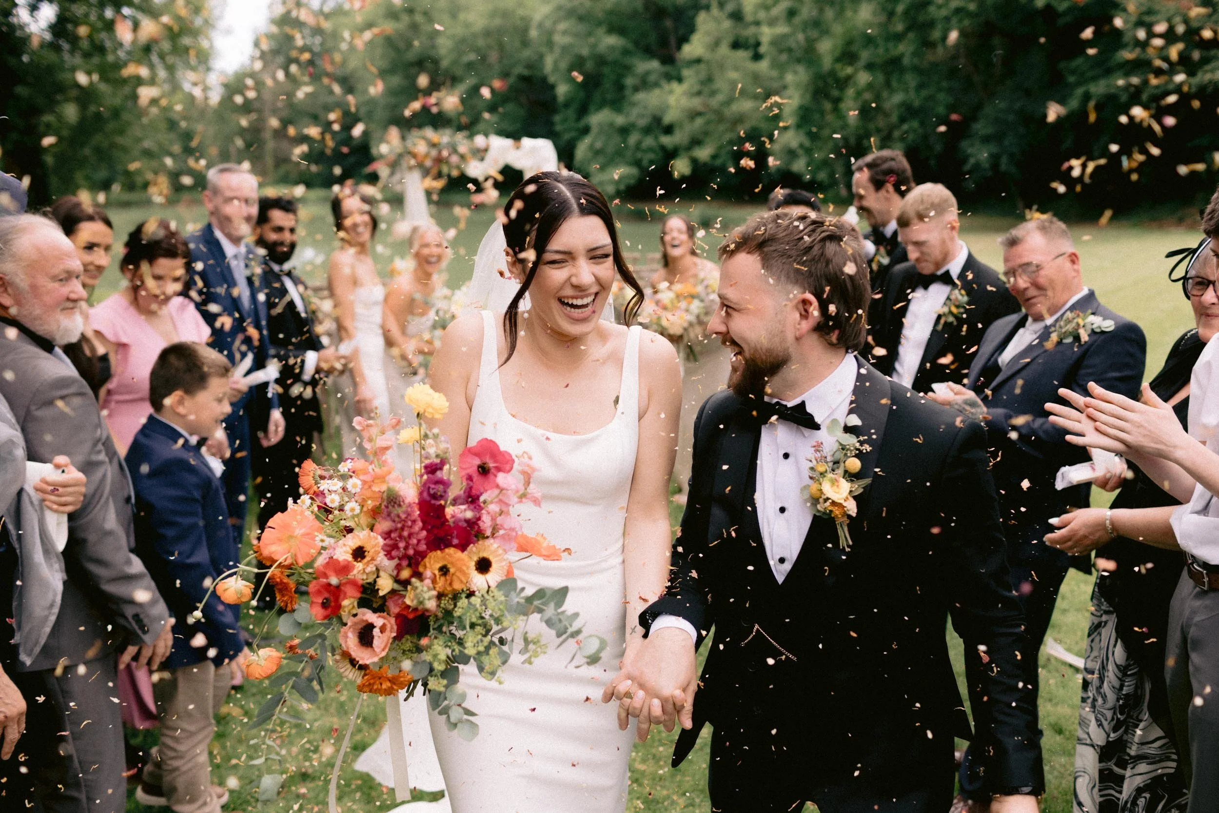 A bride and groom holding hands and smiling as they walk through a celebration with guests throwing confetti outdoors on a lush green lawn.