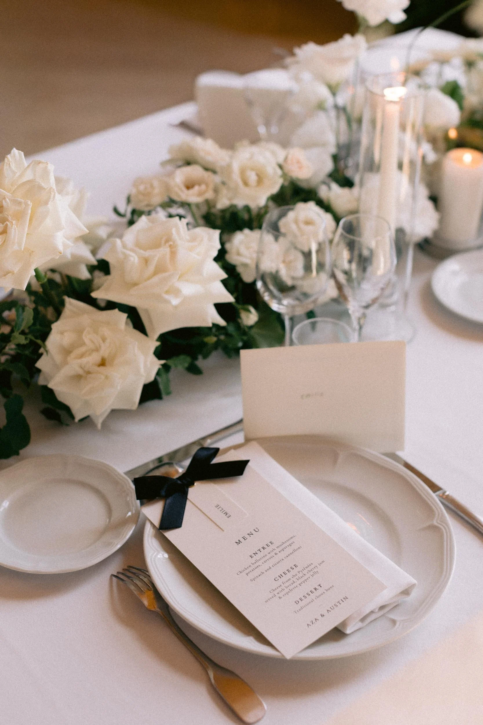 Elegant table setting with a floral centerpiece of white roses and greenery, a menu card secured with a black ribbon, white plates, silverware, wine glasses, and lit candles at a formal event.