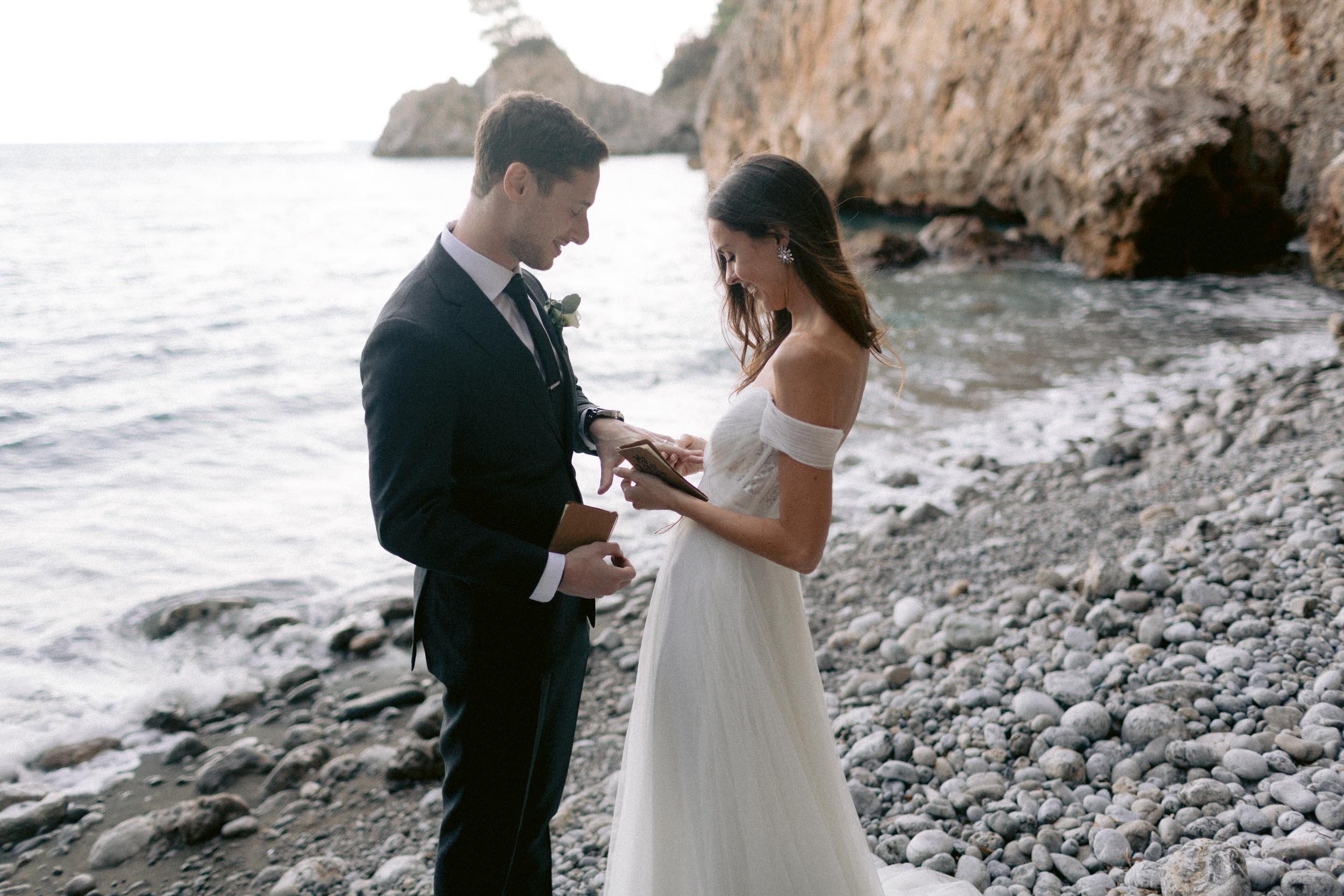 A wedding couple exchanging vows on a rocky beach near the ocean, with cliffs in the background.