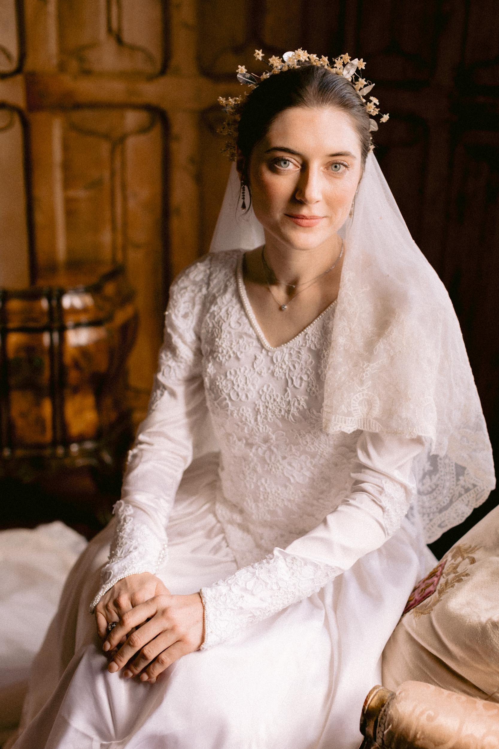 A woman in a wedding dress with lace details and a veil, sitting indoors with wooden paneling in the background, wearing a floral headpiece.