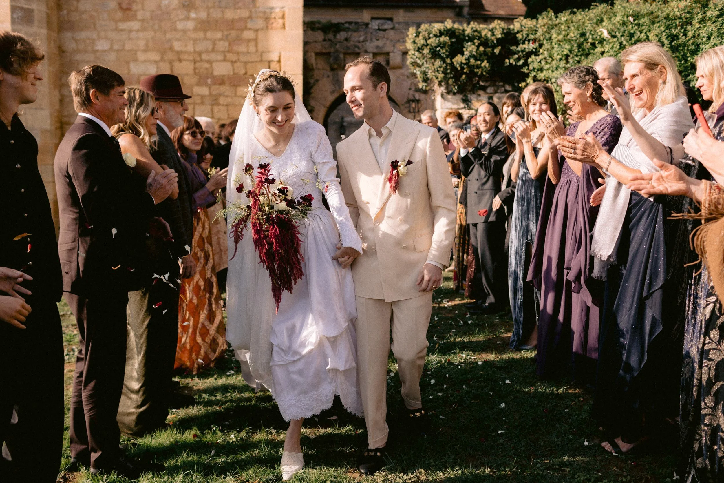 A bride and groom walking hand in hand through a crowd of wedding guests outdoors. The bride wears a white wedding dress and veil, holding a large bouquet. The groom wears a cream-colored suit. Guests cheer and applaud on both sides of the aisle, wit