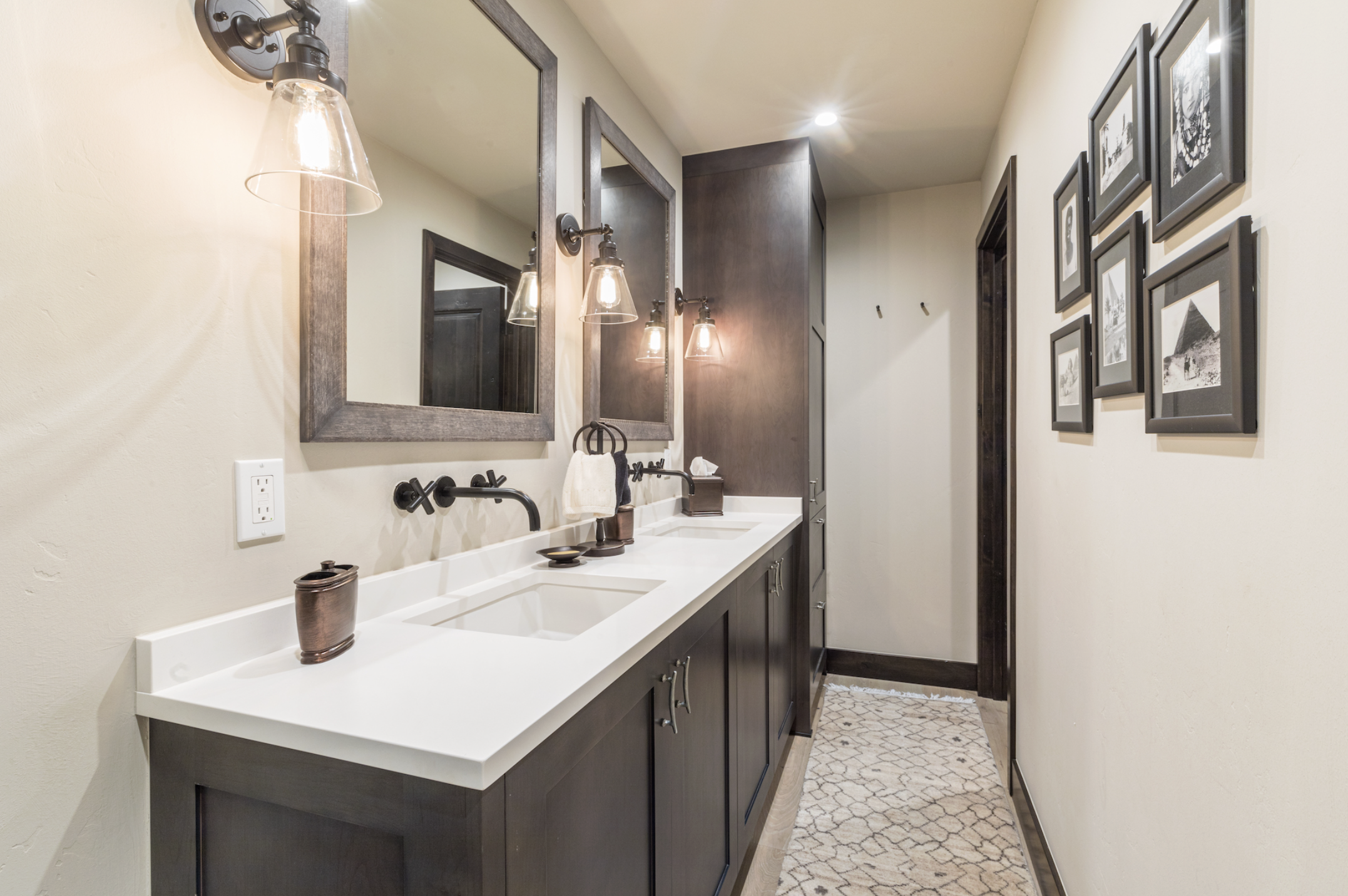 A modern bathroom with a double vanity, black framed mirrors, wall-mounted light fixtures, and a gallery of black-and-white framed photographs on the wall.