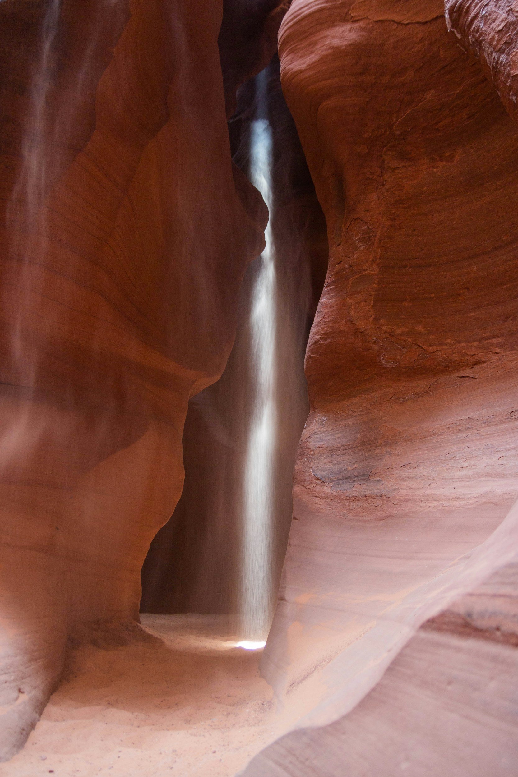 Reddish sandstone walls and a thin mist of water falling from above, with light shining at the bottom of a canyon.