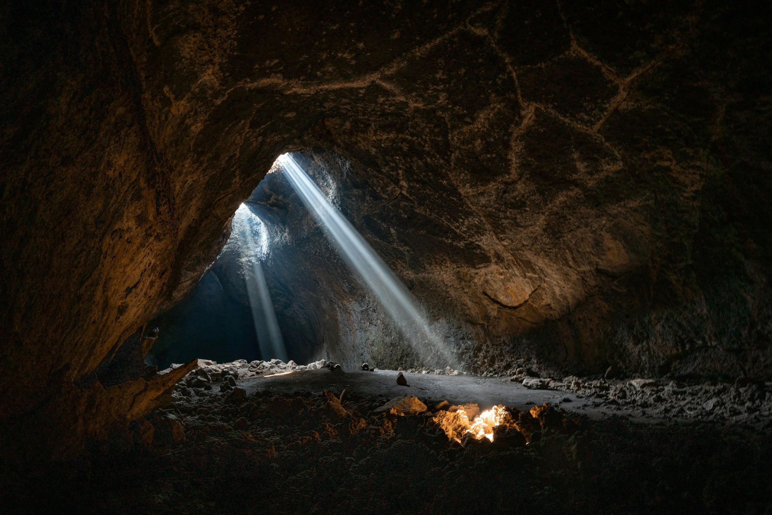 Sunlight streams through cracks in the ceiling of a dark cave, illuminating the rocky floor.