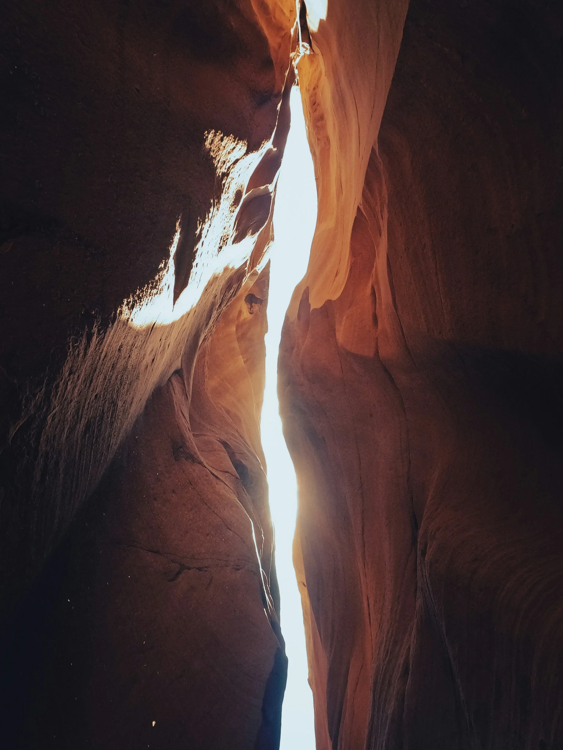 View of a thin canyon with smooth, reddish-brown rock walls and sunlight shining through the top.