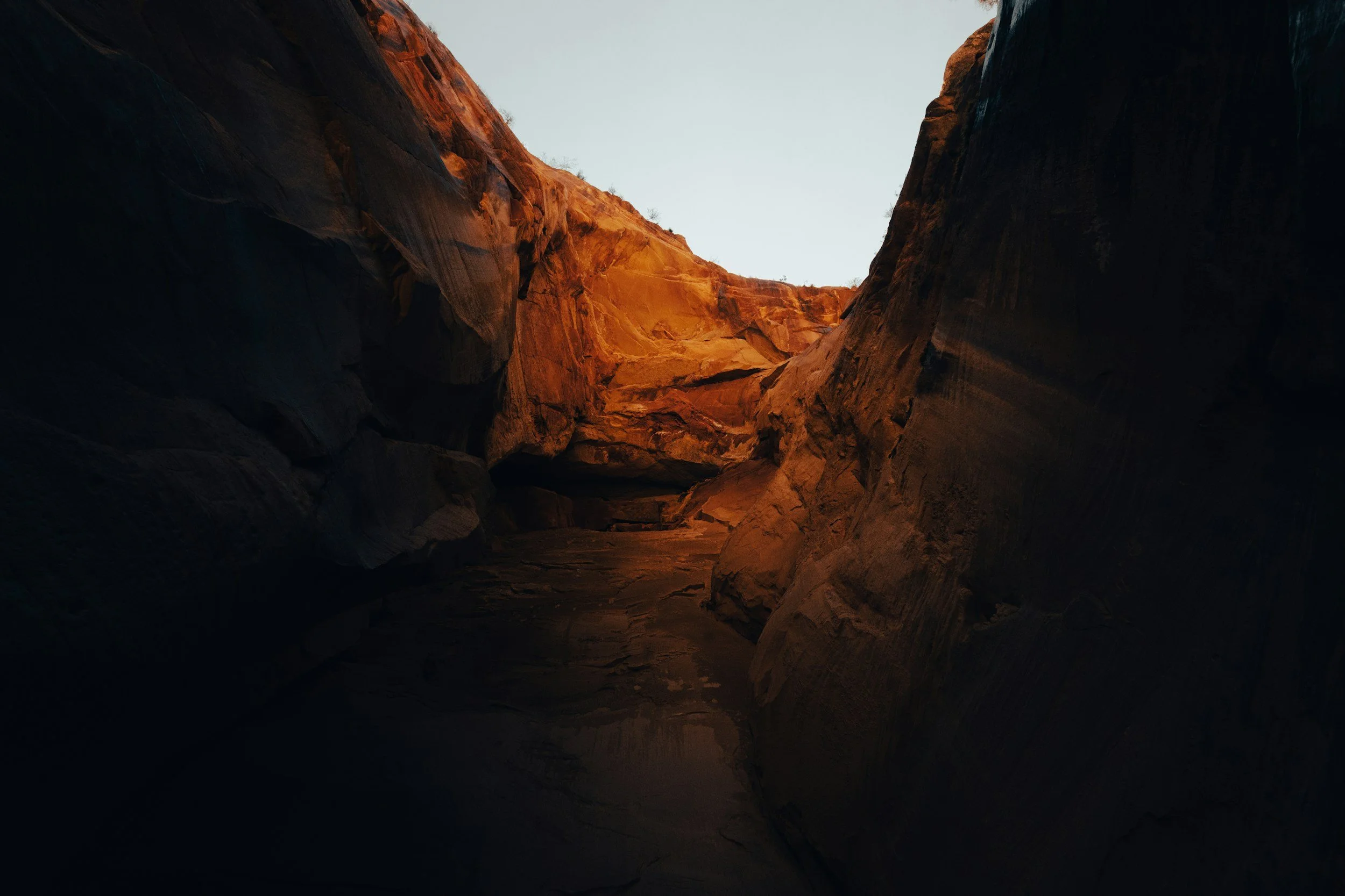 Photo of the mouth of a canyon with brown and red rock walls, illuminated by natural light from above.