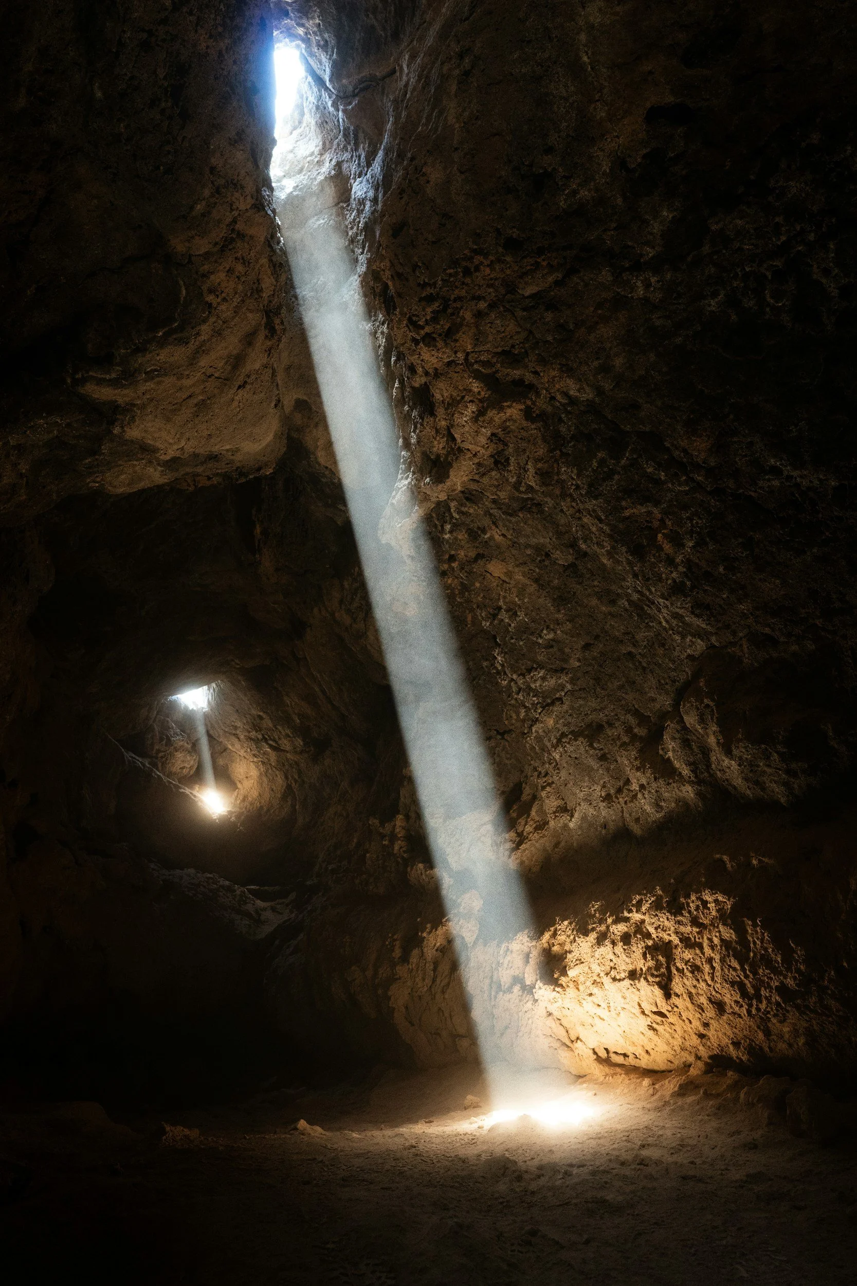 Sunlight streams through a small opening in the rocky cave ceiling, illuminating a dark cave interior.