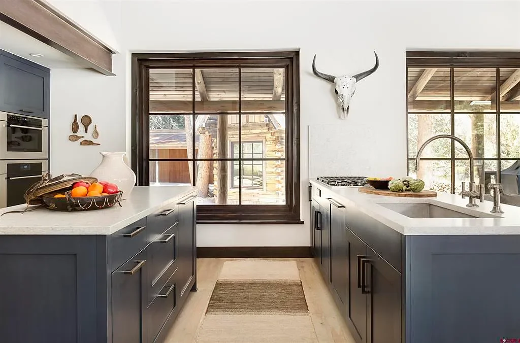 Modern kitchen with blue cabinets, white countertops, and large windows, decorated with a cow skull and a fruit basket.