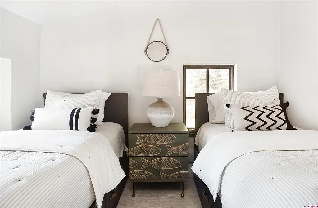 A bedroom with two twin beds, white bedding, and black headboards. A nightstand with a fish design is between the beds, holding a white lamp. There is a small window behind the nightstand and a circular mirror hanging above it.