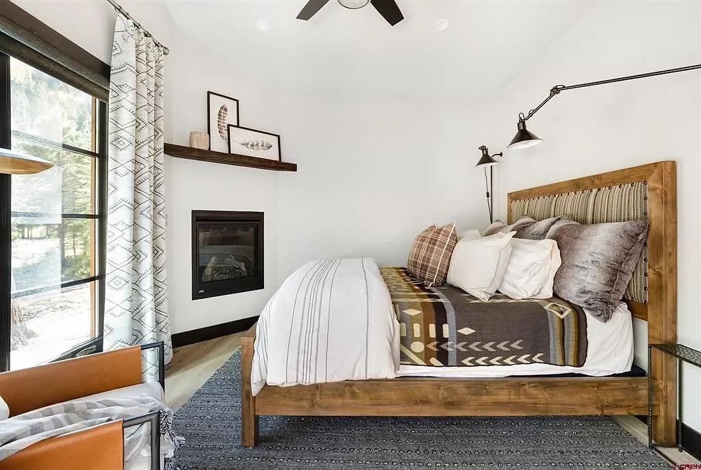 A cozy bedroom with a large wooden bed, multiple pillows, and a Southwestern style blanket. The room has a fireplace, a large window with patterned curtains, and wall-mounted reading lamps. Decor includes framed artwork on a shelf above the fireplace and a patterned rug.