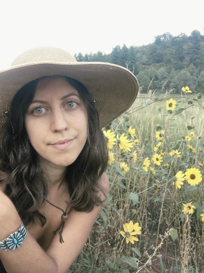 Young woman with long wavy dark hair, wearing a wide-brimmed straw hat and a beaded bracelet, taking a selfie in a field of yellow flowers with a hillside and trees in the background.