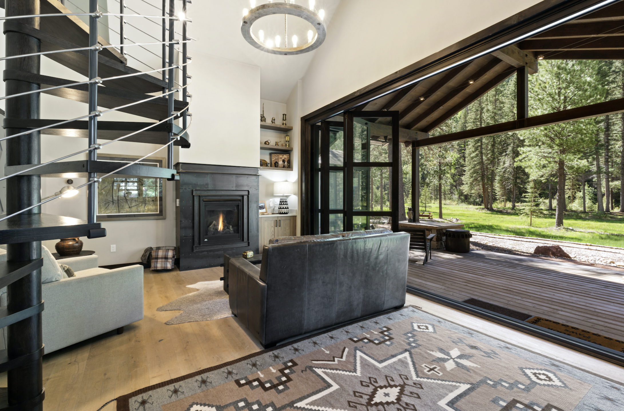 Living room with a fireplace, sofa, and a sliding glass door leading to a wooded outdoor area. Modern decor with shelves, a lamp, and a patterned rug.
