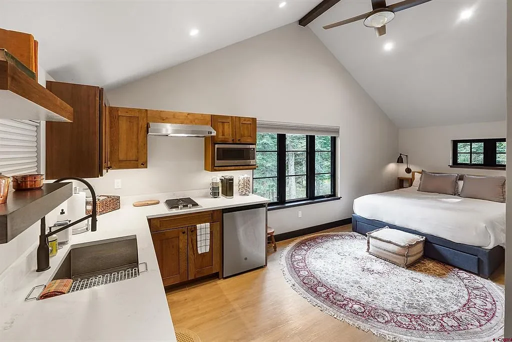 A cozy studio apartment with a small kitchen area and a bed, featuring a large window with trees outside, a ceiling fan, and a round patterned rug.