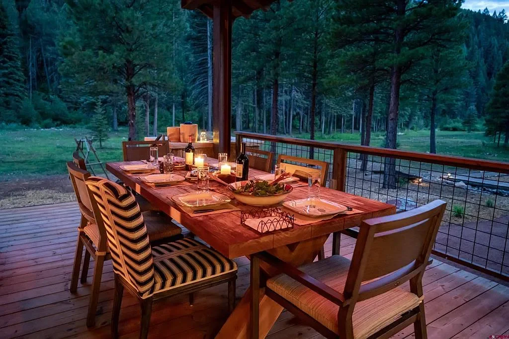 Outdoor dining table set on a wooden deck, illuminated by candles, with a forested background.