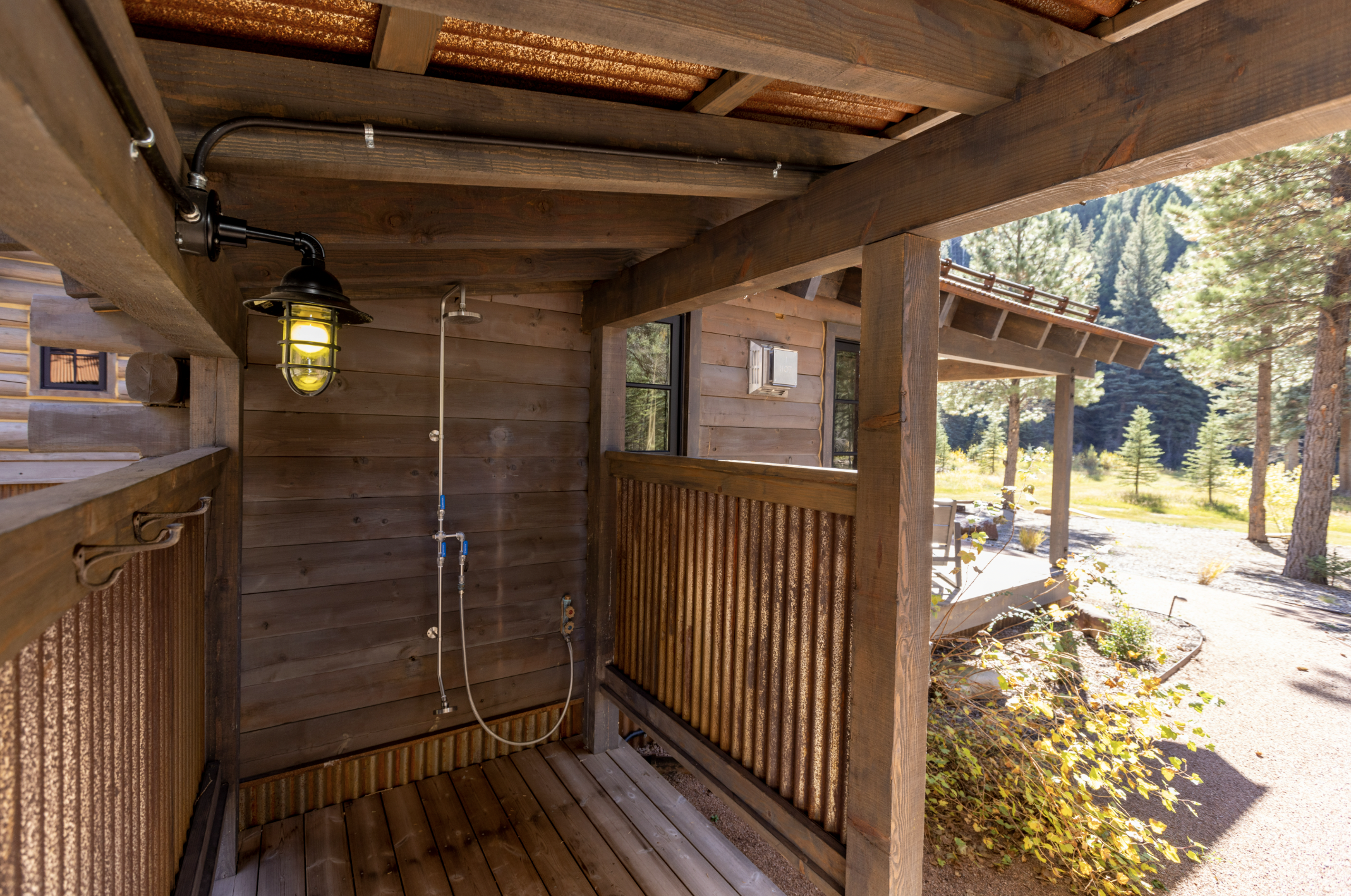 An outdoor shower area with a wooden structure, mounted black shower head with a flexible hose, and a warm-colored wooden railing, overlooking a forested landscape with trees and mountains in the background.