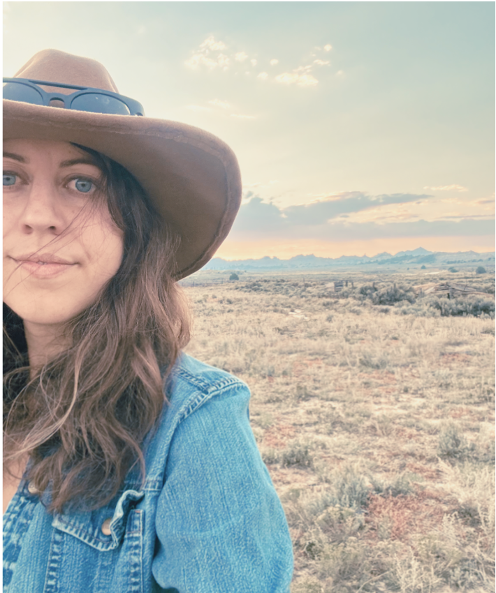 A woman wearing a wide-brimmed hat, sunglasses on top, and a denim jacket standing in a vast, open desert landscape during sunset or sunrise.