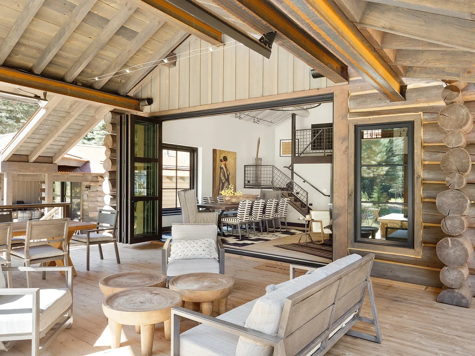A spacious rustic living area with wooden walls and ceiling beams, large windows, and modern furniture, including armchairs, a dining table with striped chairs, and decorative logs on the wall.