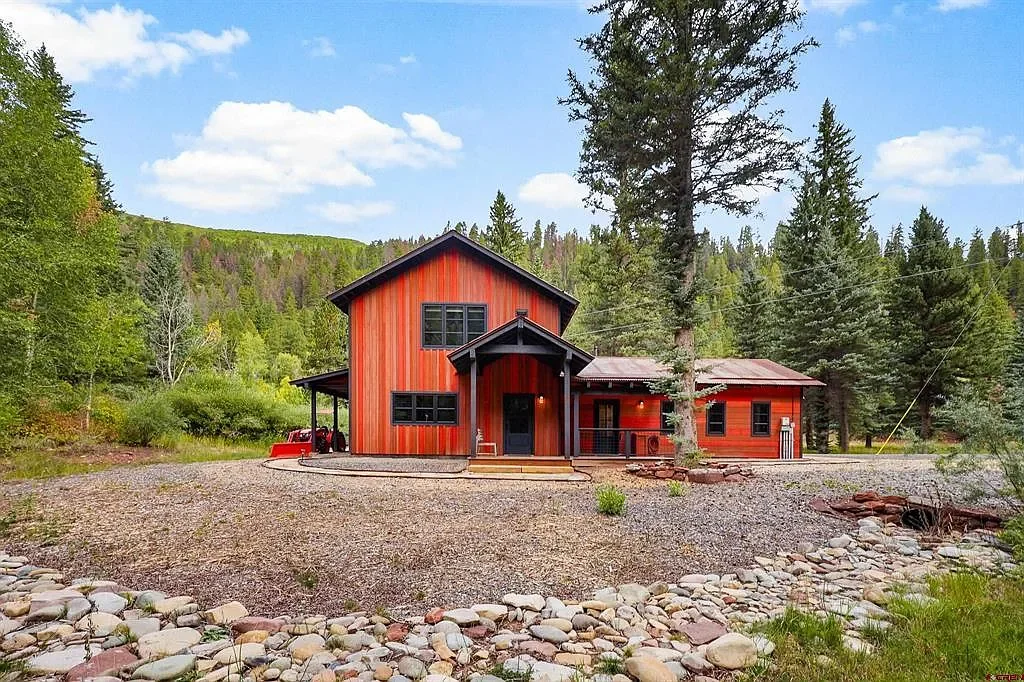 Red wooden house surrounded by trees and mountains with a gravel yard in the foreground.