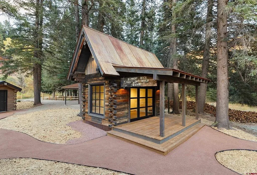 Small rustic cabin with a metal roof and large windows, surrounded by tall trees, with a wooden porch and a pathway nearby.