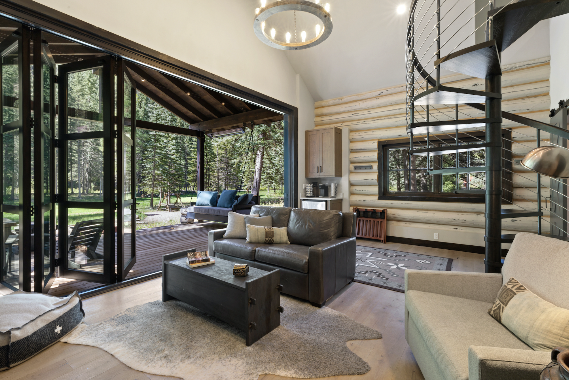 Living room with large glass windows and door leading to outdoor patio, featuring a sofa, armchairs, coffee table, and spiral staircase, with natural wood and modern decor.