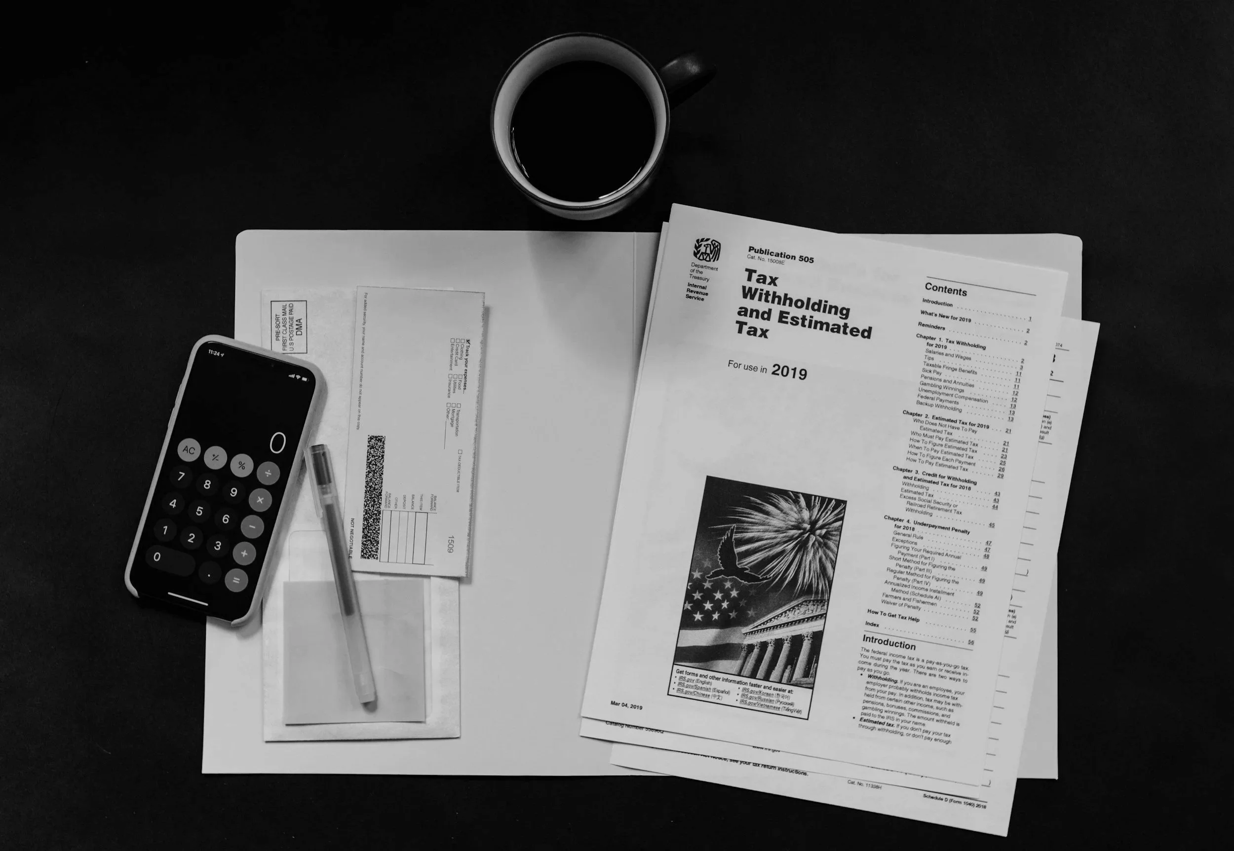 Black desk with a cup of coffee, tax documents, a calculator, pen, and envelope lay on top of a white mat.